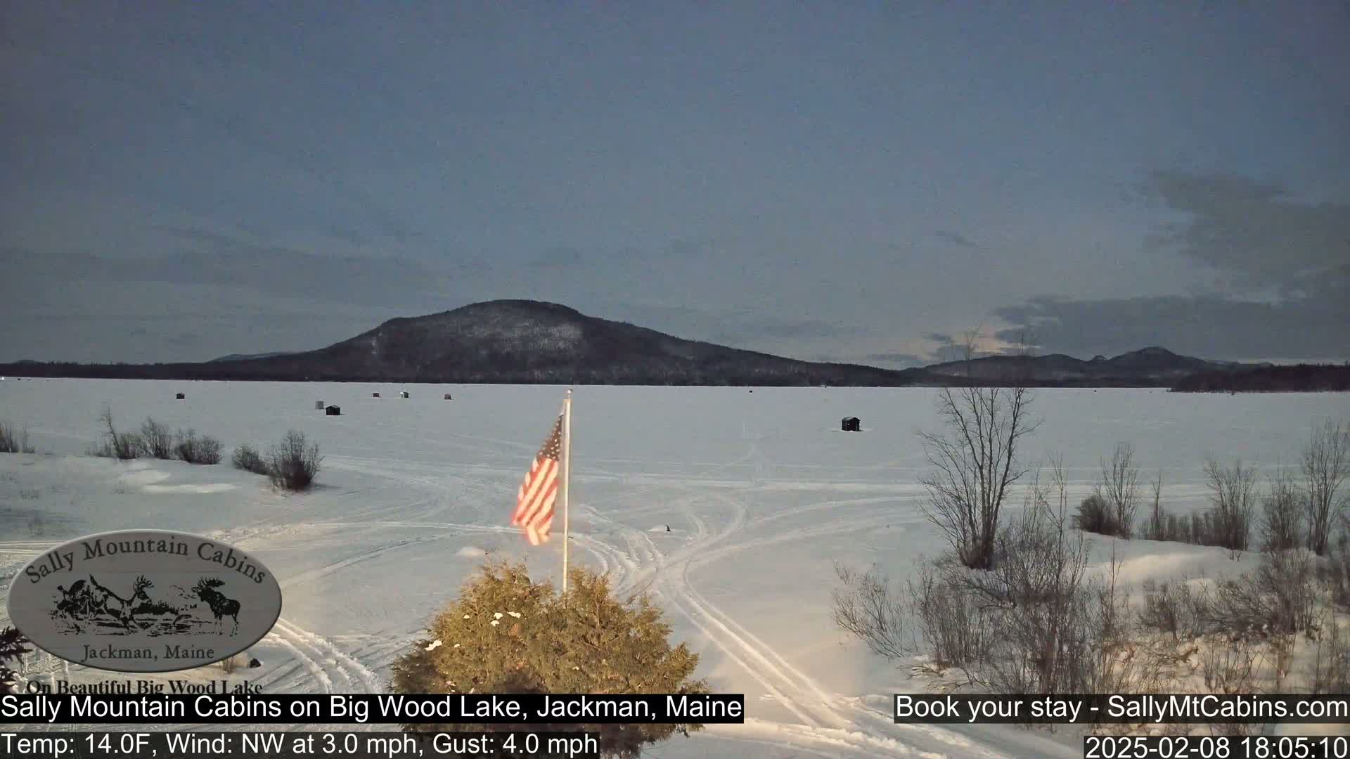 A frozen lake under a twilight sky, with a snow-covered shoreline, a few ice fishing shelters, and an American flag; a mountain range is visible in the background.
