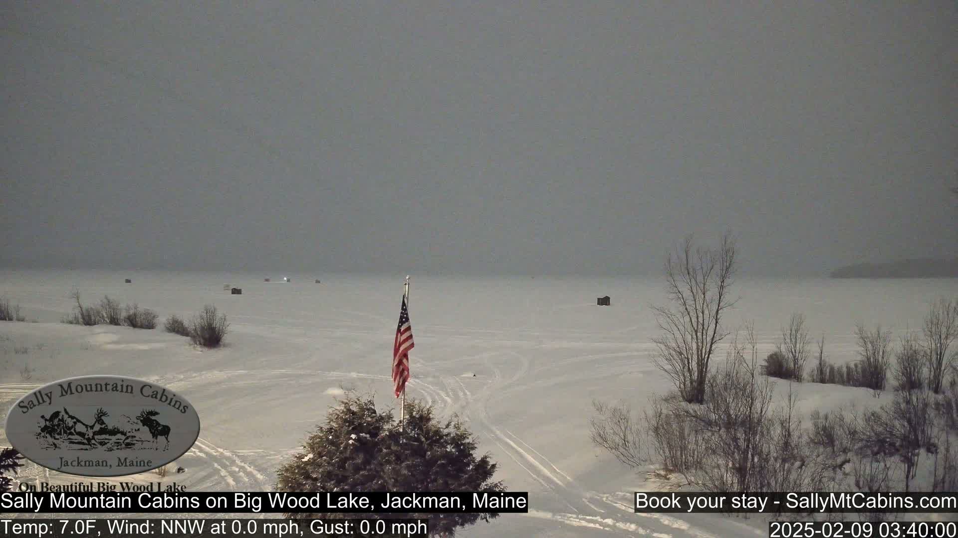A snow-covered frozen lake is visible under an overcast sky, with a small American flag planted near the shore and some ice fishing shelters scattered across the lake's surface.