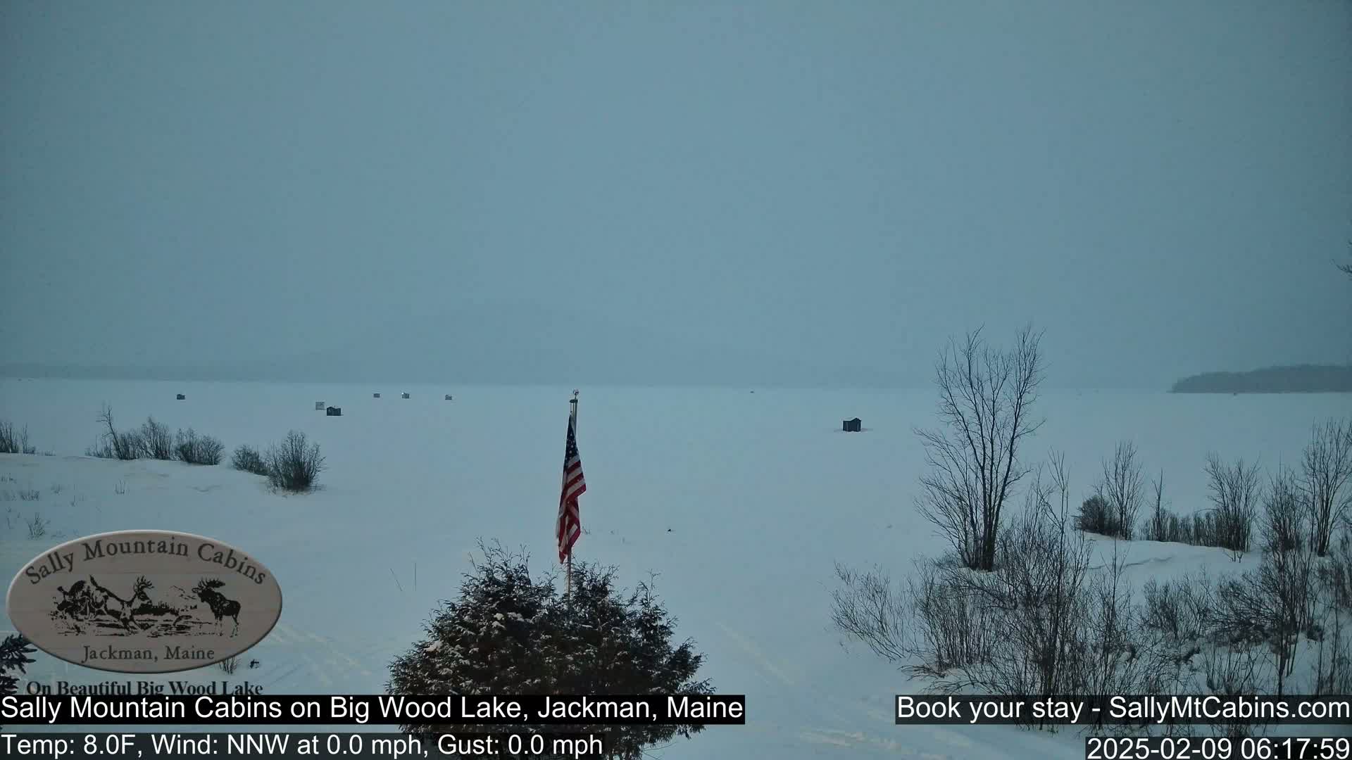 A snow-covered frozen lake with several small structures and an American flag, under an overcast sky.