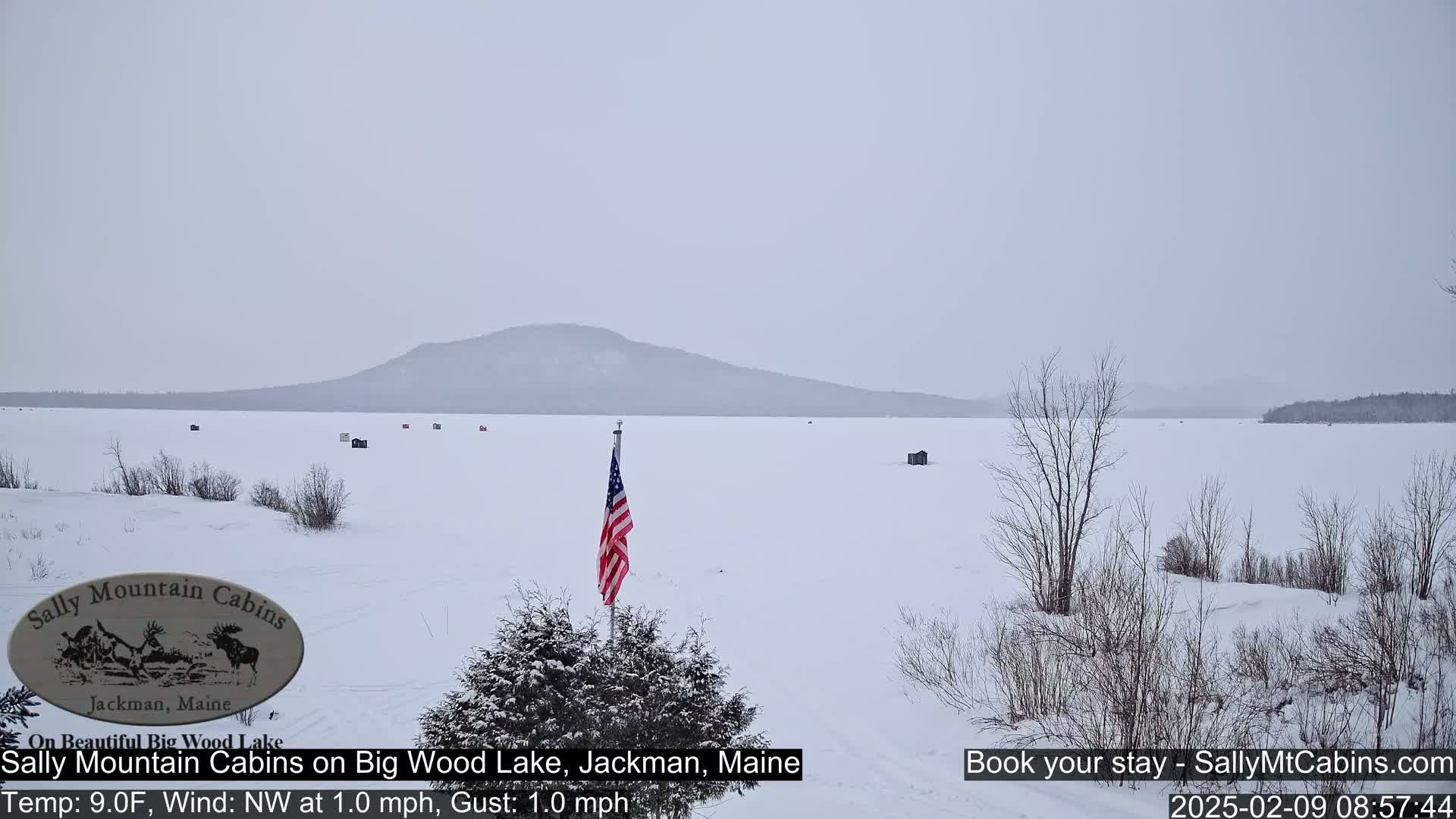 A snow-covered frozen lake stretches to a distant, snow-dusted mountain range under an overcast sky, with a few ice fishing huts scattered across the lake and an American flag planted near the foreground.