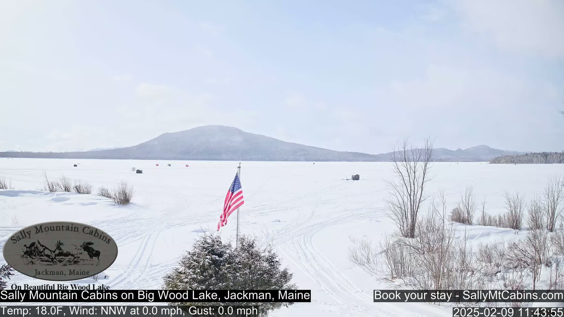 A frozen lake, dotted with a few small structures, stretches towards snow-covered mountains under a bright, clear sky.