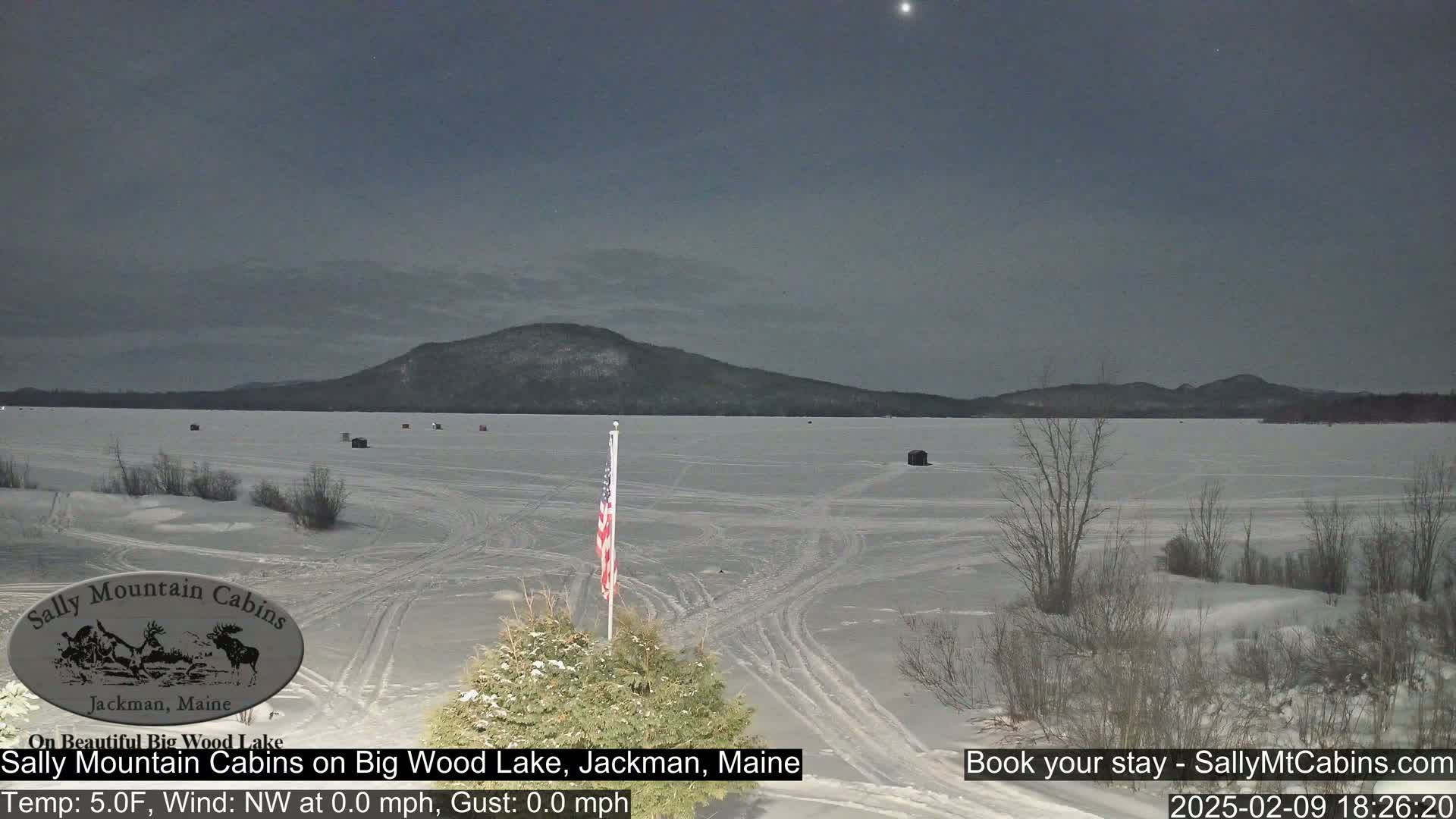 A frozen lake stretches toward snow-covered mountains under a twilight sky, with an American flag visible in the foreground.
