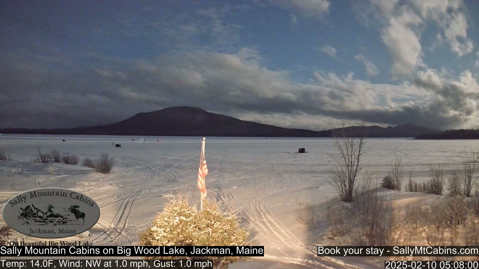 A frozen lake under a partly cloudy night sky, with a small American flag and snow-covered vegetation in the foreground, and distant mountains under a moonlit sky.