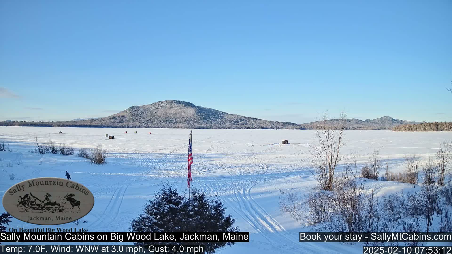 A snow-covered frozen lake stretches to a wooded mountain range under a clear blue sky.