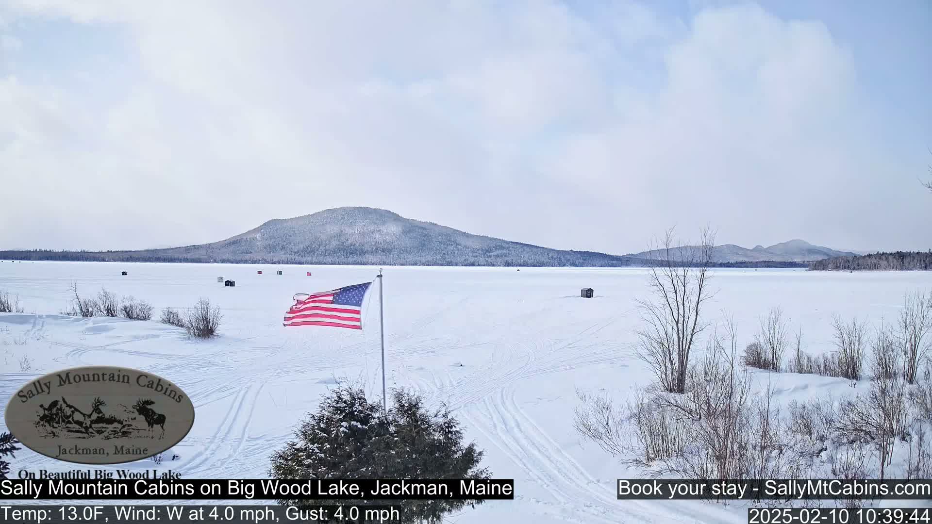 A frozen lake stretches to snow-covered mountains under a mostly clear sky, with an American flag waving near the foreground.