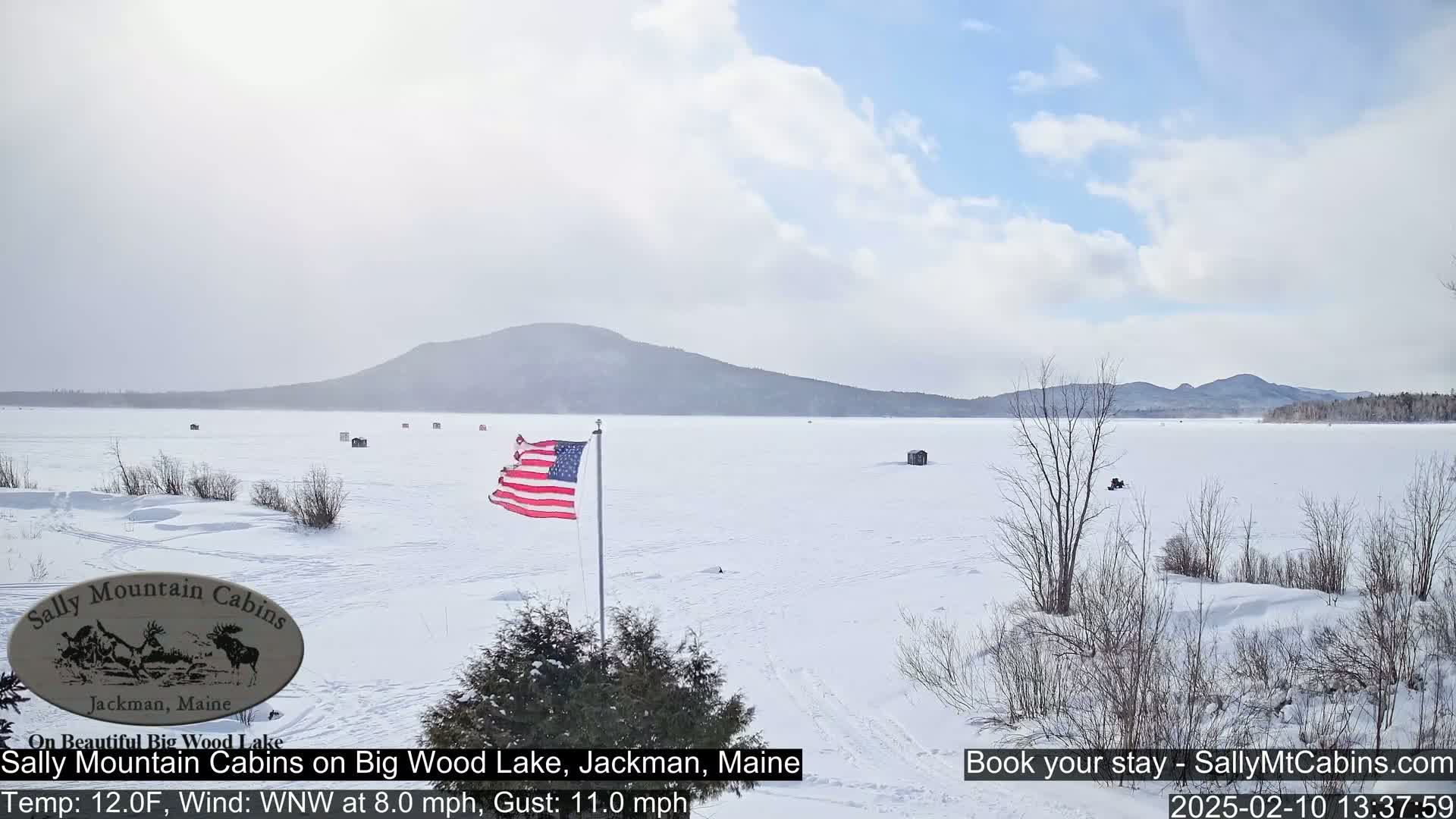 A snow-covered frozen lake stretches to a distant mountain range under a partly cloudy sky, with a few ice fishing shacks scattered across the ice and an American flag waving in the breeze.