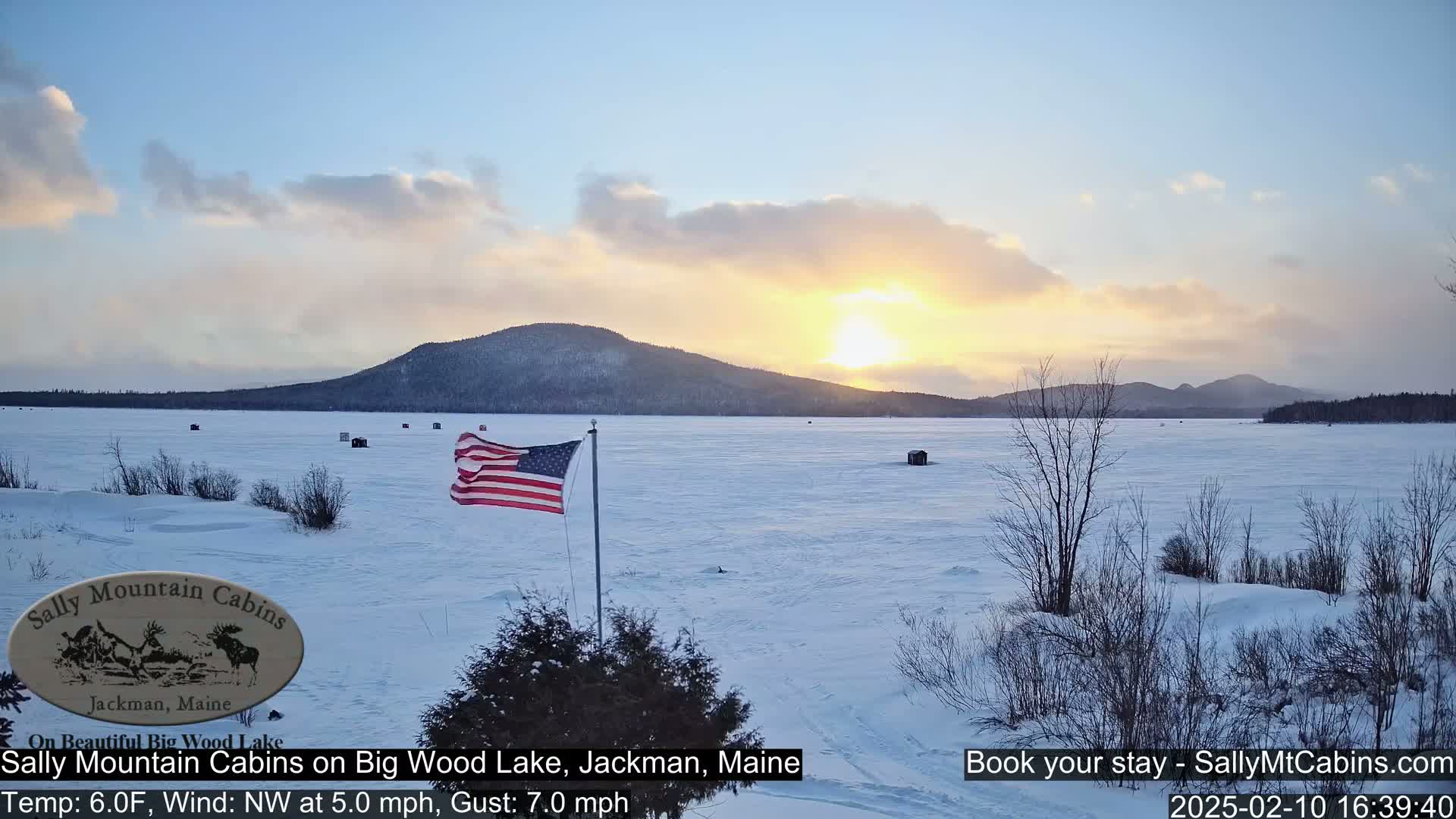 A frozen lake at sunset, under a partly cloudy sky, with a few ice fishing shacks and an American flag visible.