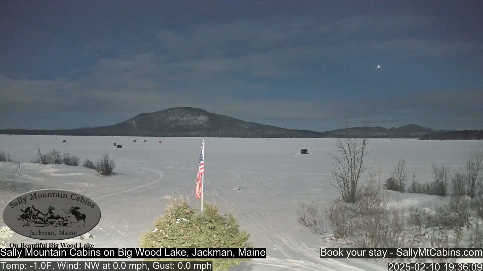 A frozen lake stretches towards a snow-covered mountain under a clear night sky, with a small American flag planted in the foreground.