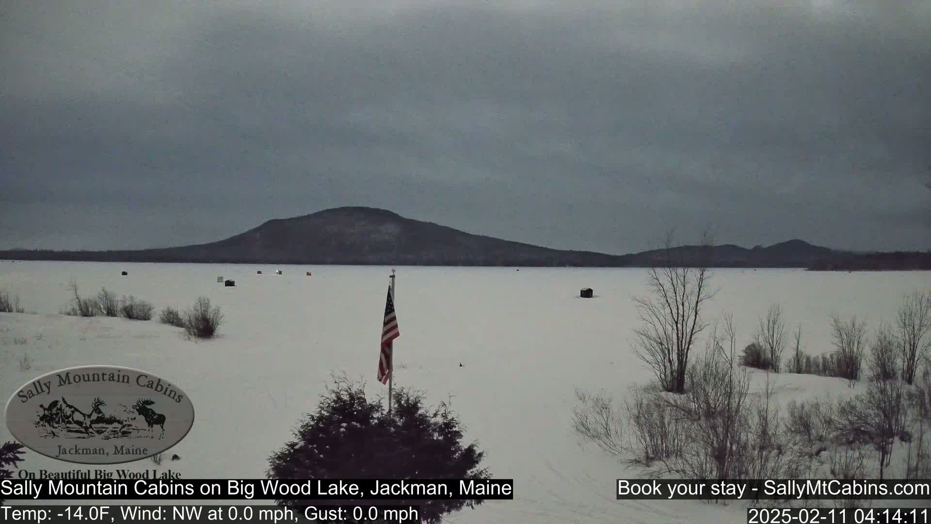 A snow-covered frozen lake stretches out before a dark mountain range under an overcast sky, with a few ice fishing shelters scattered across the ice and an American flag in the foreground.