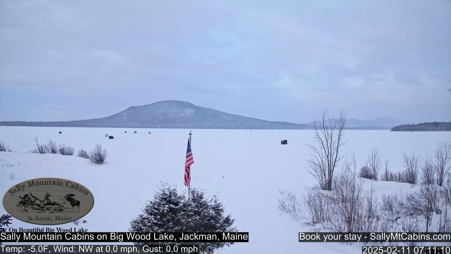 A snow-covered frozen lake stretches to a distant mountain range under a cloudy sky, with a few ice fishing huts scattered across the ice and an American flag planted near the foreground.