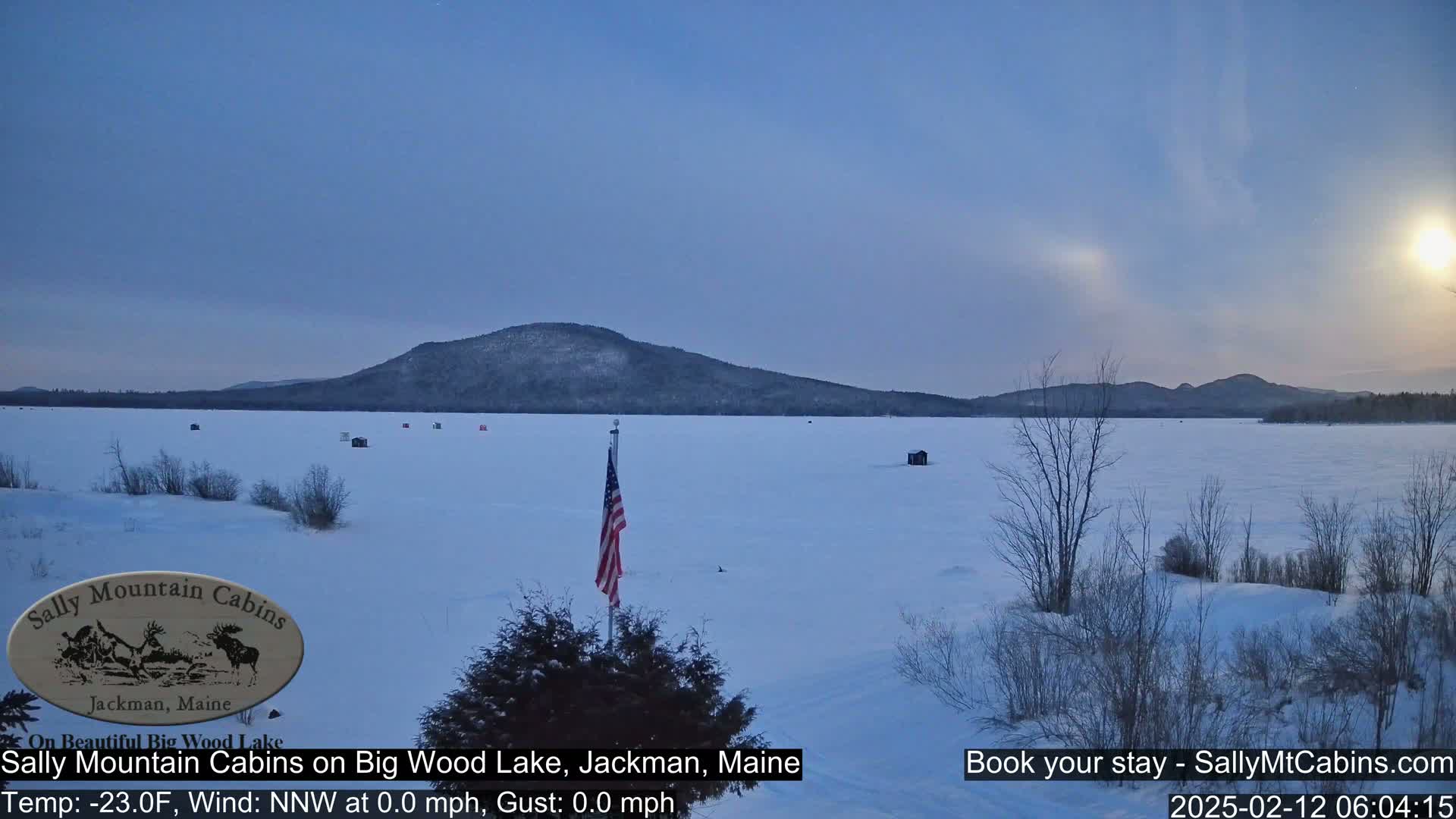 A frozen lake with several ice fishing huts is visible under a twilight sky, with a snow-covered shoreline and distant mountains.