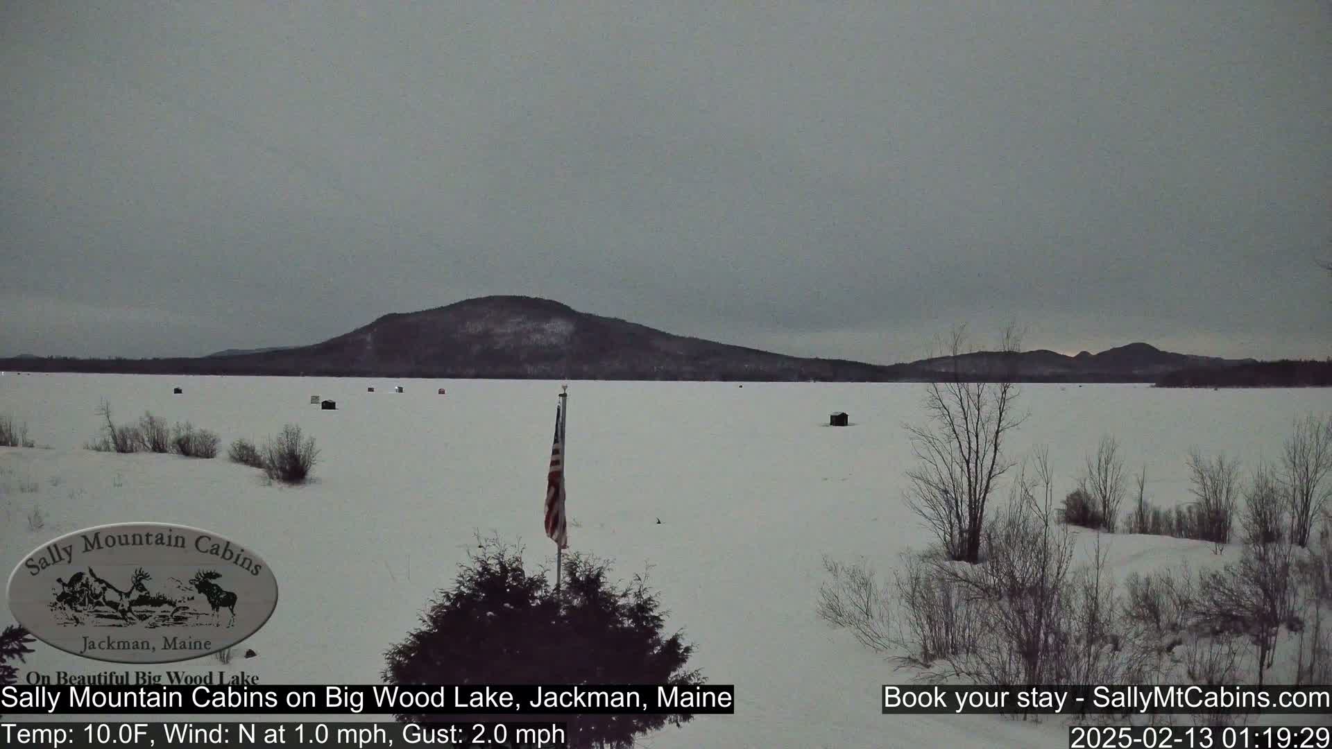 A frozen lake with scattered ice fishing shacks, a distant mountain range, and a small American flag are visible under an overcast sky.