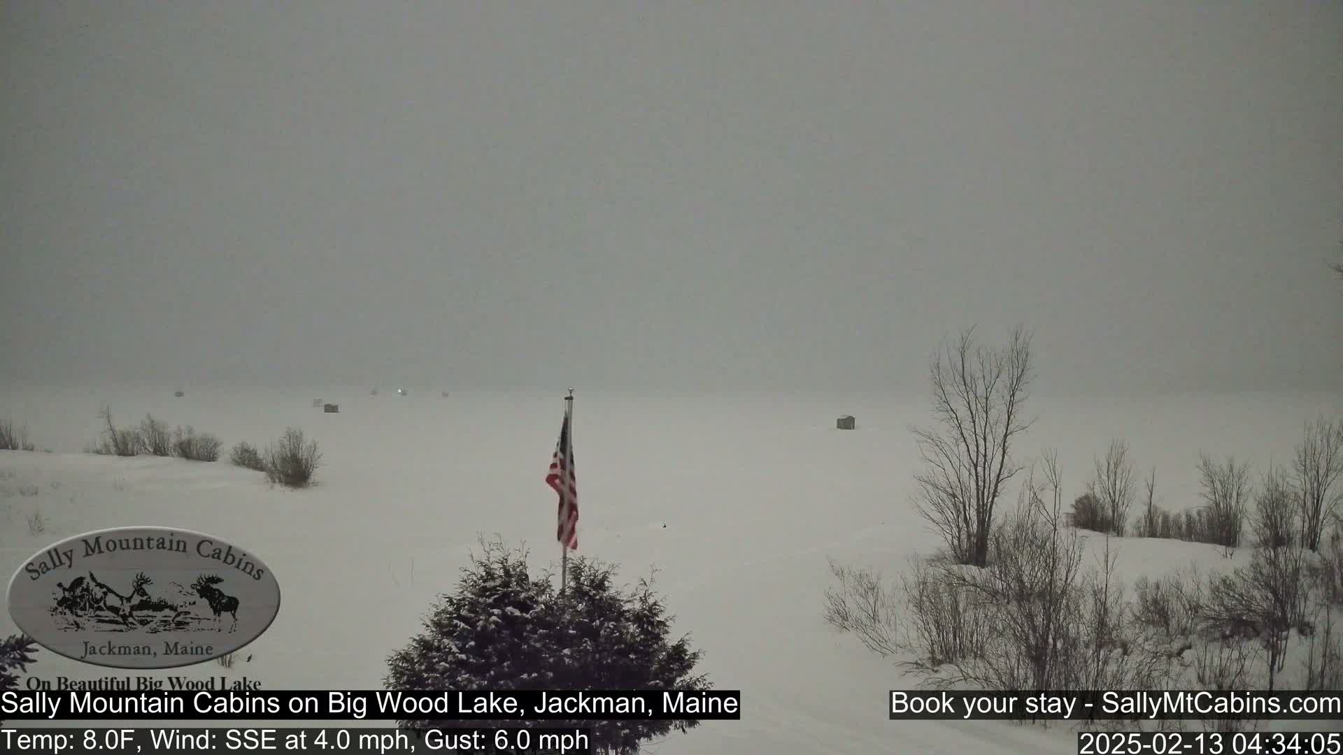 A snow-covered landscape shows a frozen lake with a few structures visible in the distance under a gray, overcast sky.