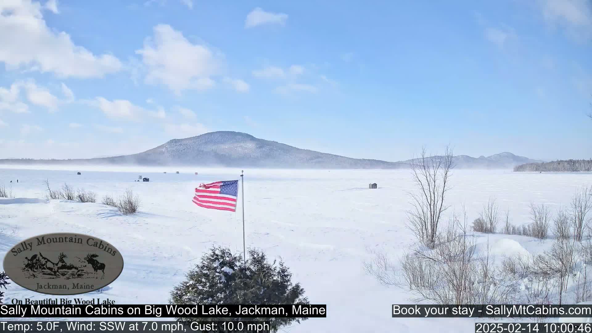 A snow-covered frozen lake stretches to a distant mountain range under a mostly sunny sky with a light wind.