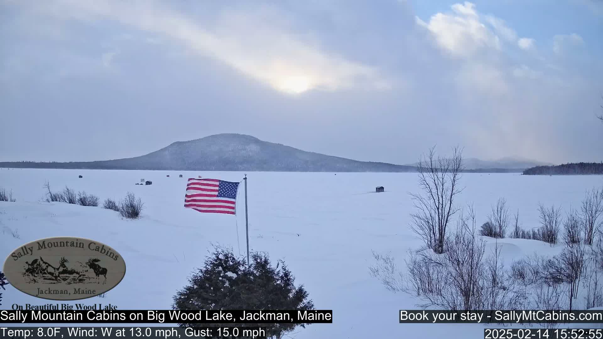 A snow-covered frozen lake stretches to a distant mountain under a mostly cloudy sky, with an American flag flying near the foreground.