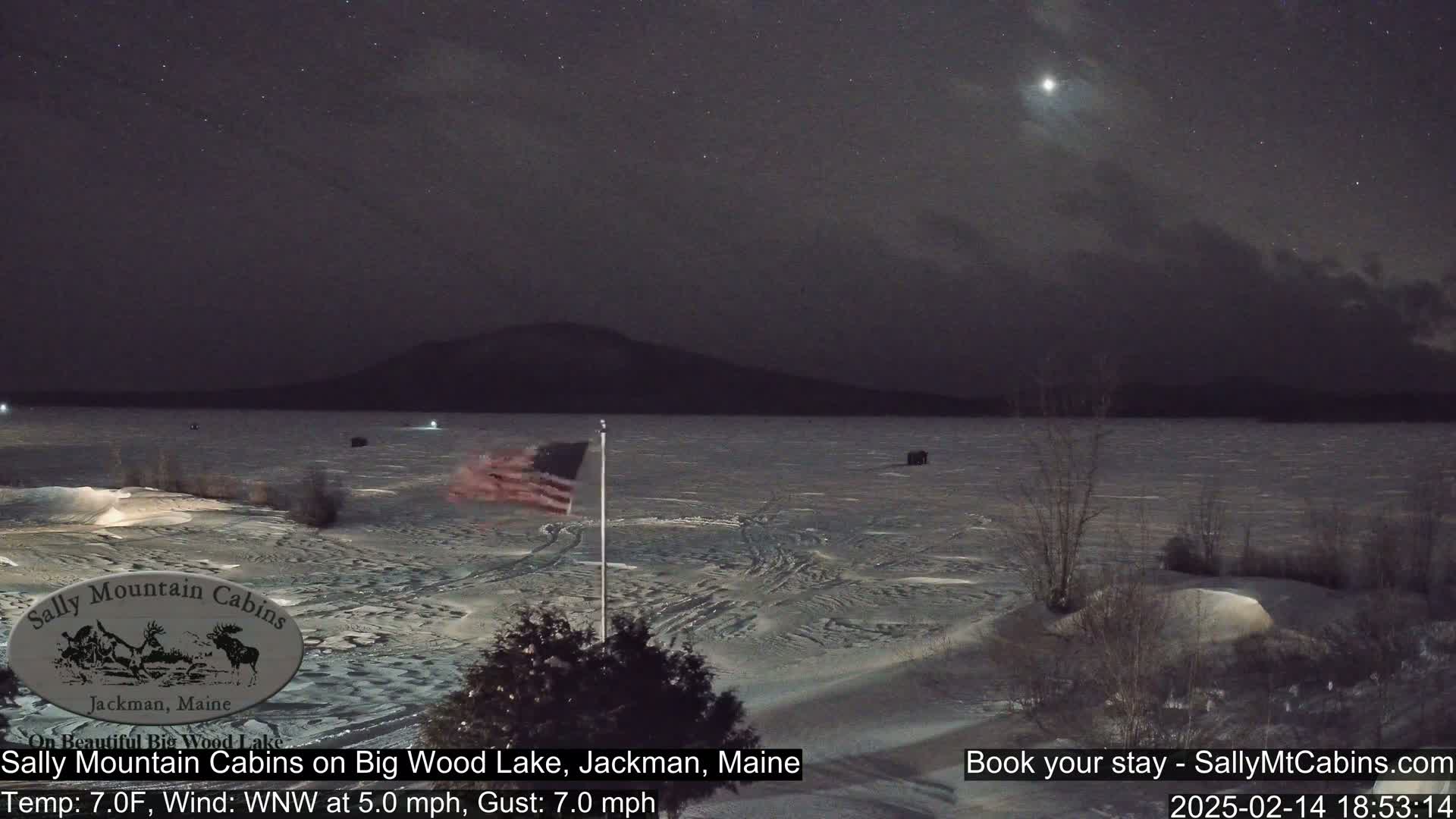 A snow-covered frozen lake is visible under a partly cloudy night sky with a bright star or planet, a distant mountain range, and an American flag on a pole in the foreground.