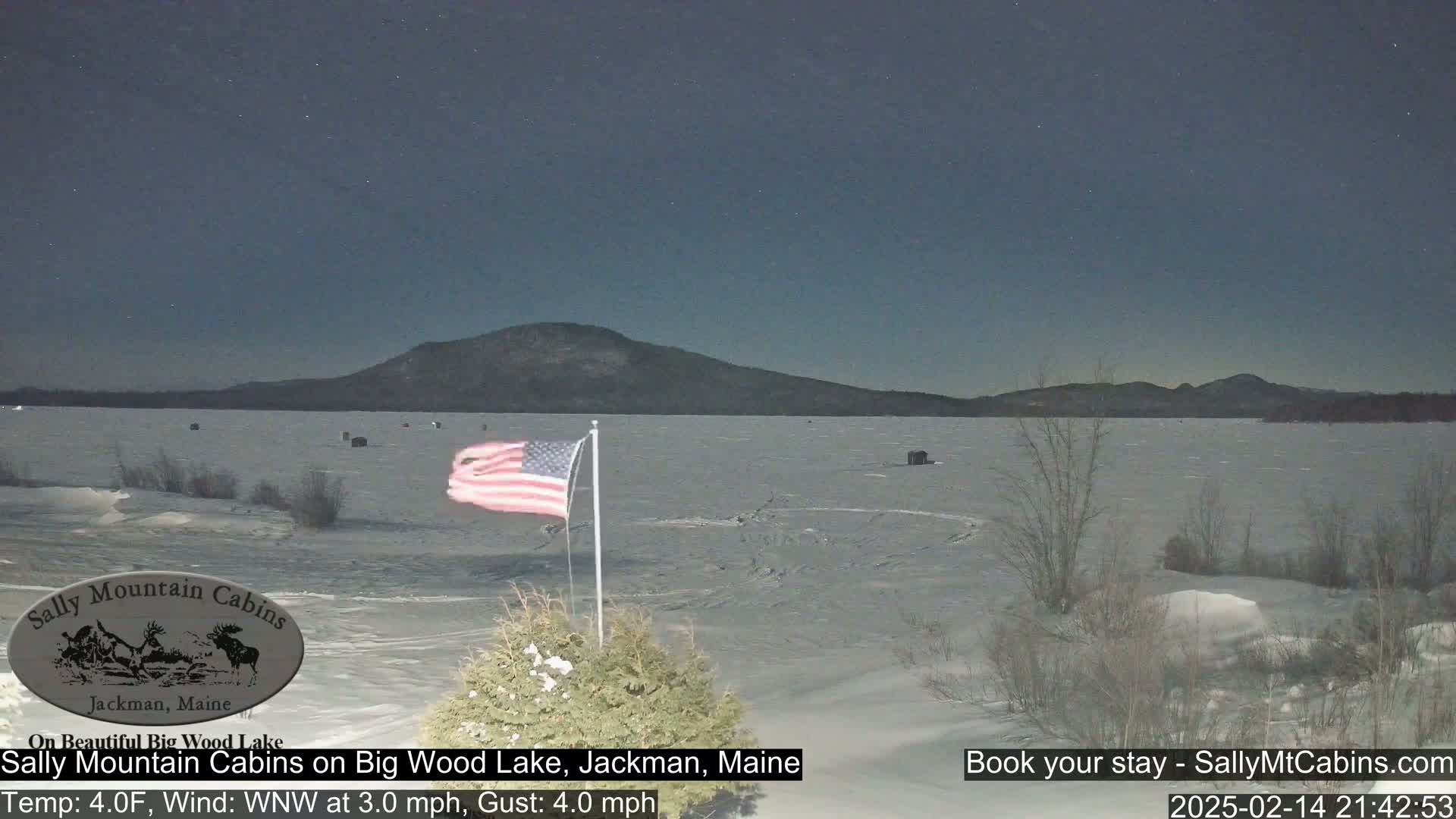 A frozen lake stretches out under a clear night sky, with a distant mountain range visible in the background and an American flag waving in the foreground.