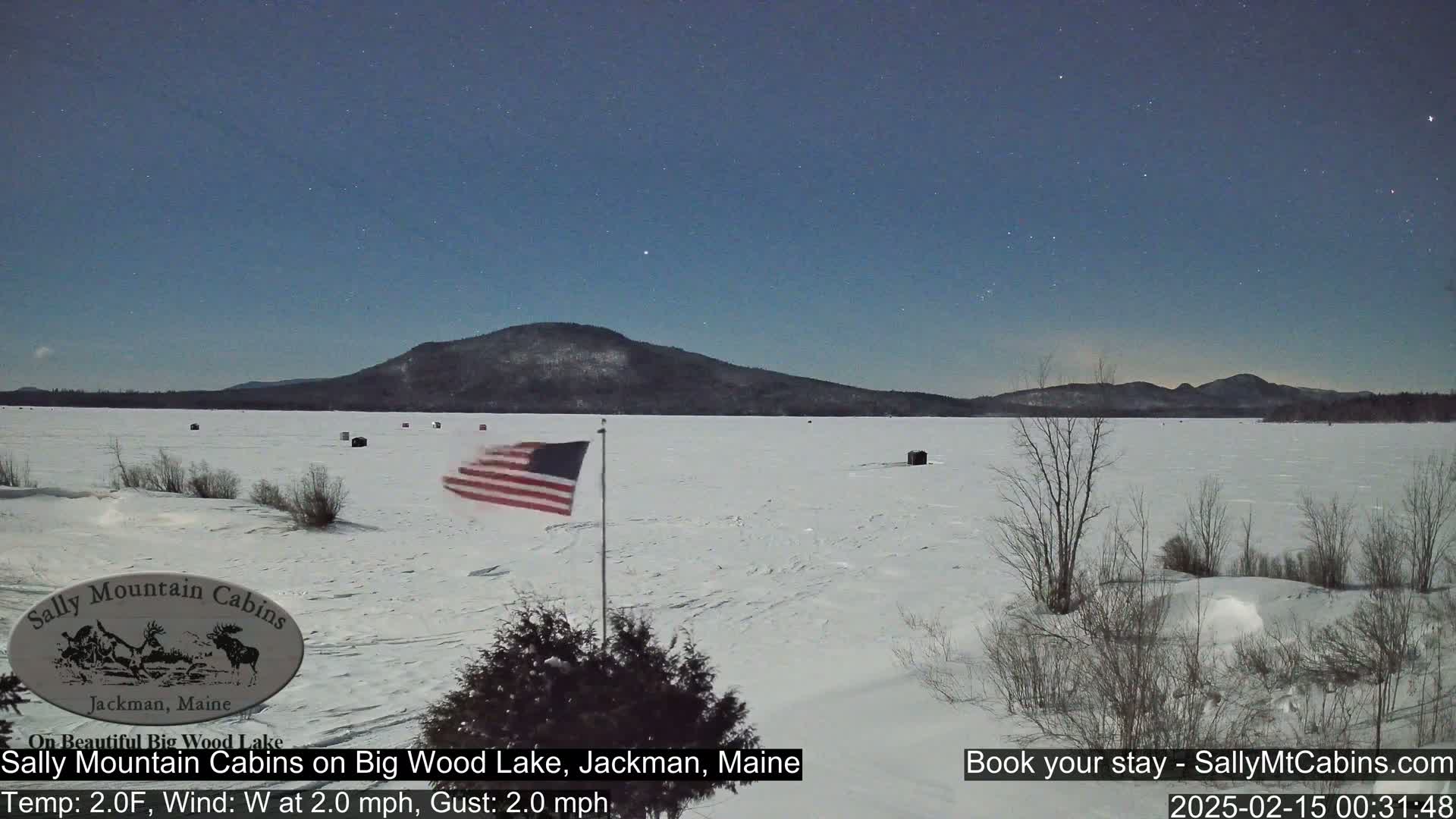 A snow-covered frozen lake stretches out under a clear, starry night sky, with a distant mountain range and a small American flag waving in the light breeze.