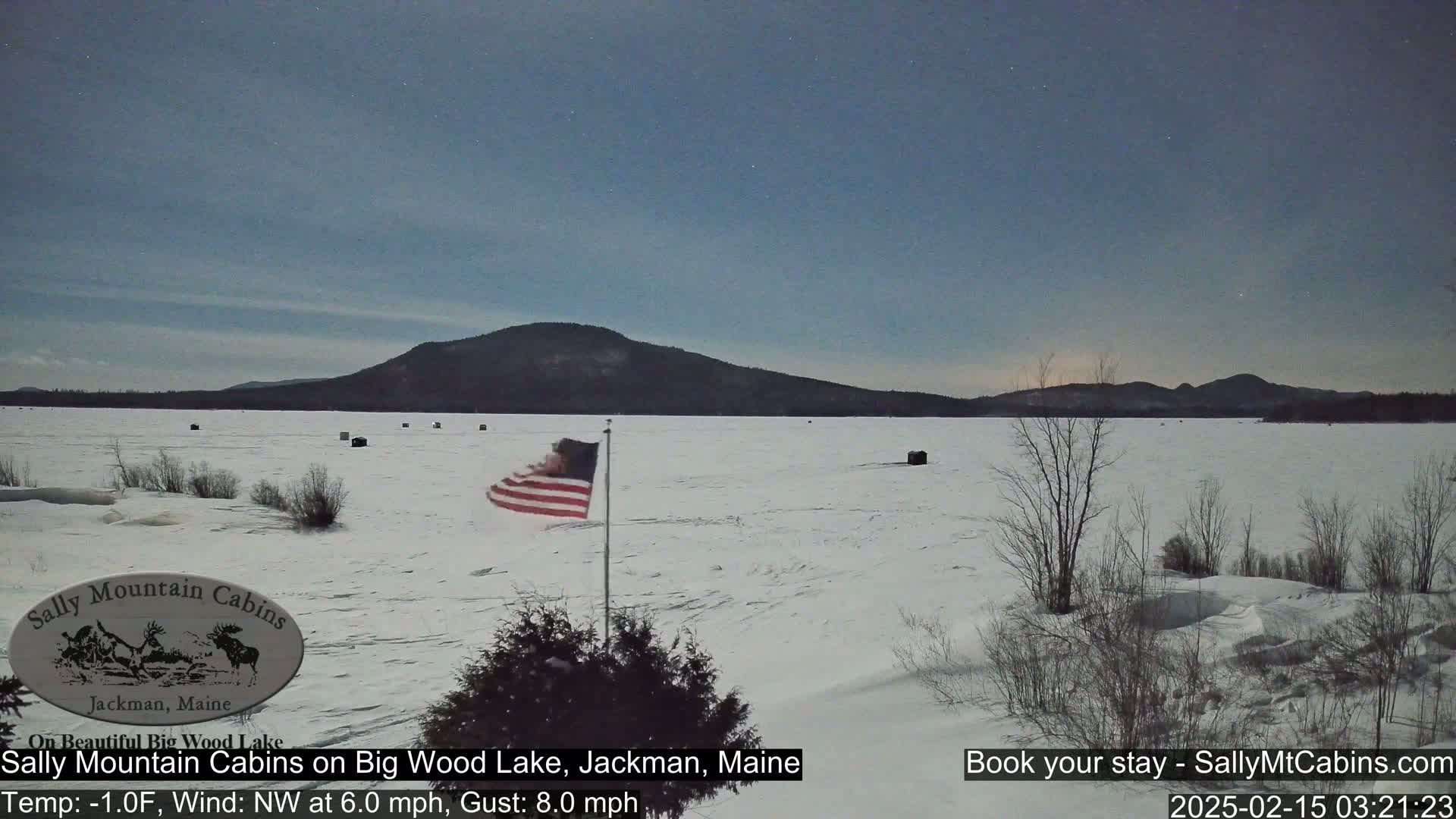 A snow-covered frozen lake stretches to a distant mountain range under a twilight sky, with a flag waving in a northwest wind.