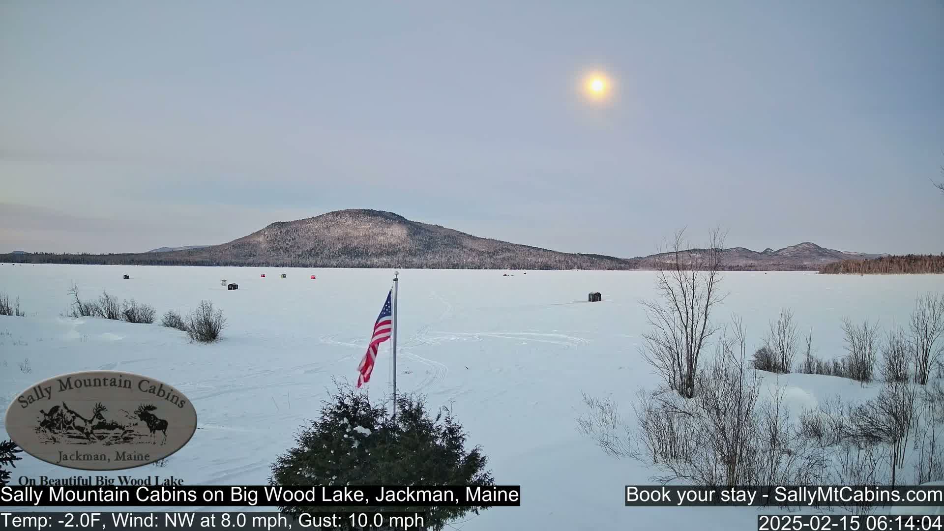 A frozen lake stretches to snow-covered mountains under a pale sun in a cold, windy winter scene.