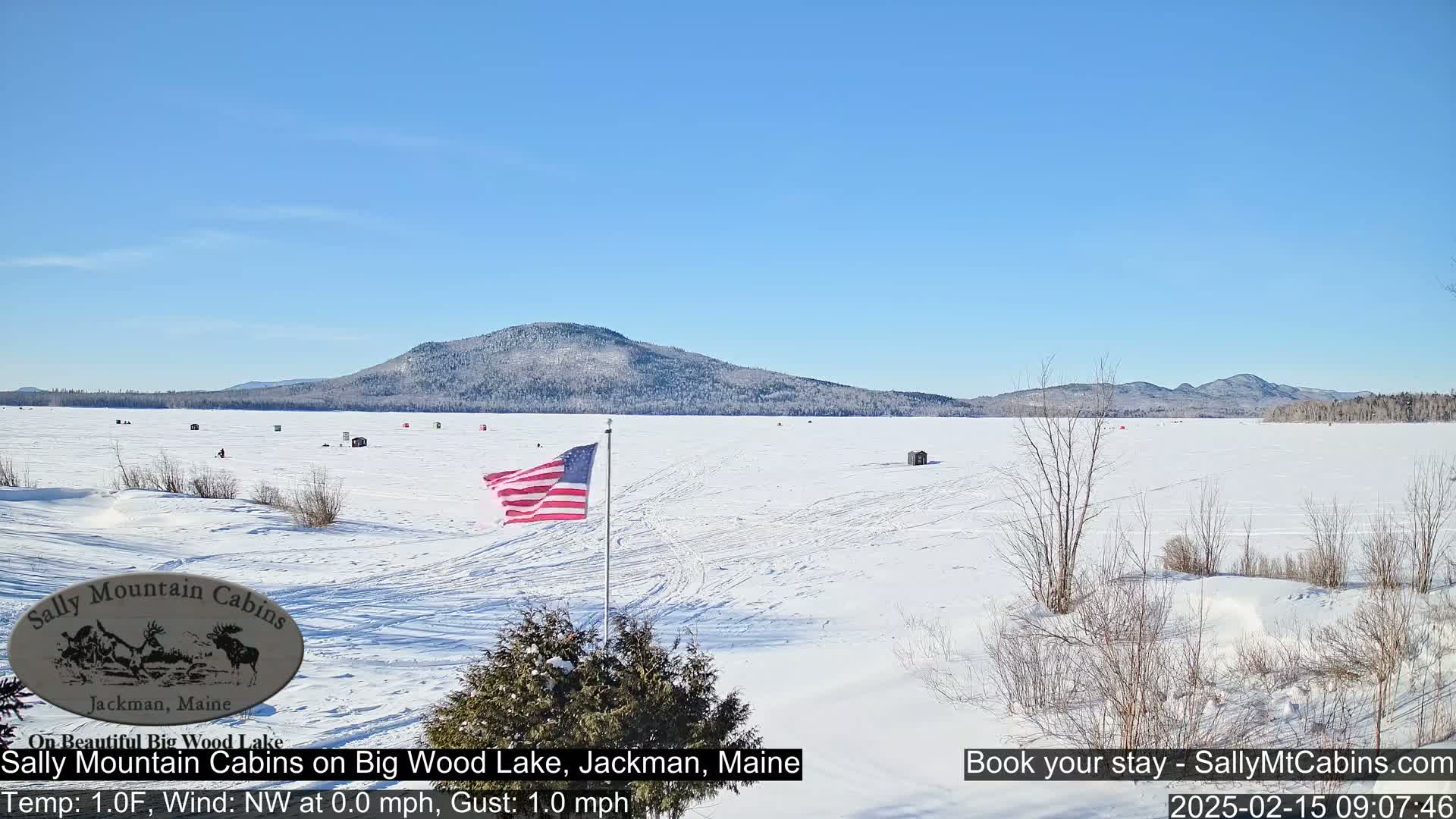 A frozen lake with scattered ice fishing shelters stretches out beneath a clear blue sky, backed by snow-covered hills.