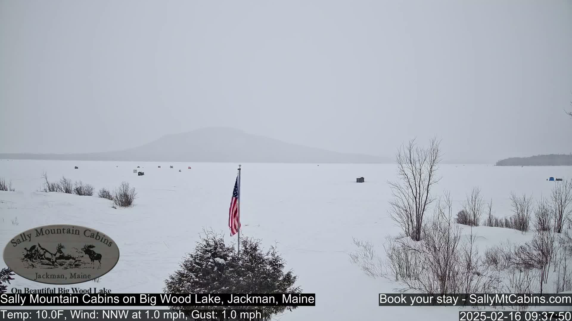 A snow-covered frozen lake with several ice fishing huts and a distant mountain range is visible under an overcast sky.