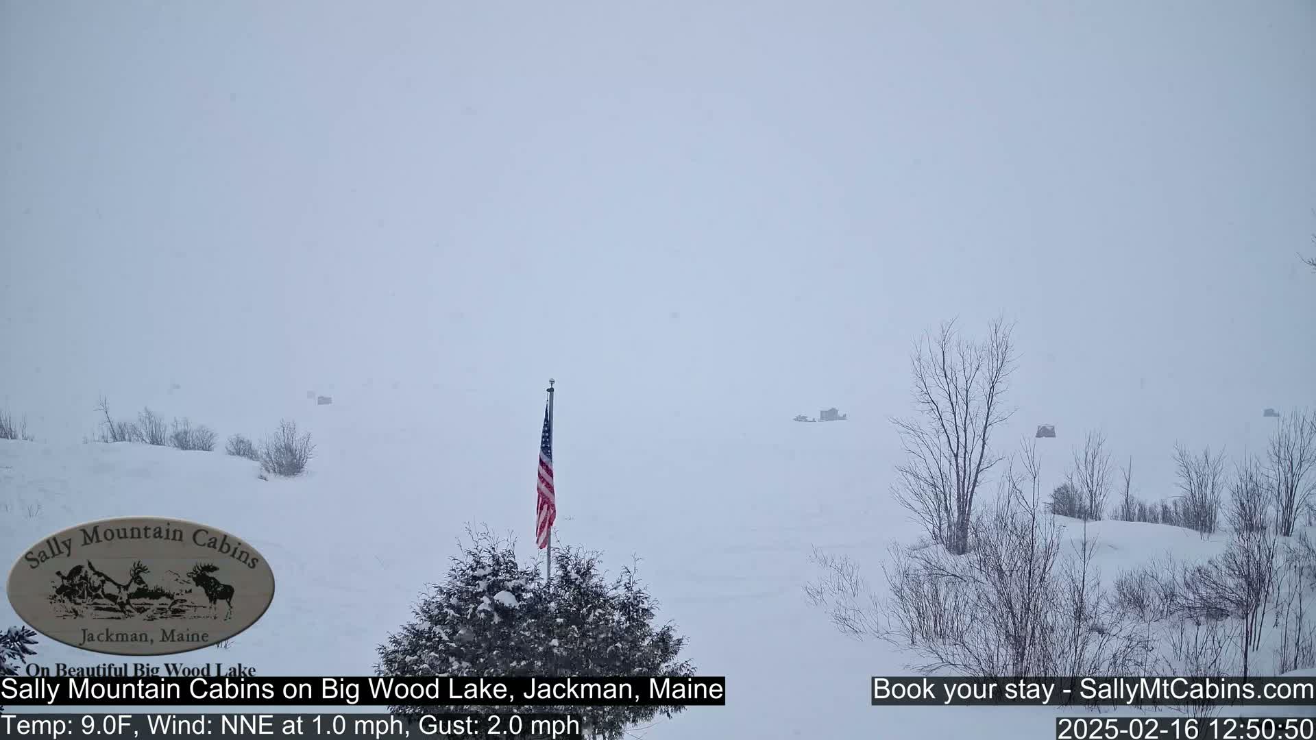 A snow-covered landscape with several small structures visible in the distance under a heavy, gray sky.