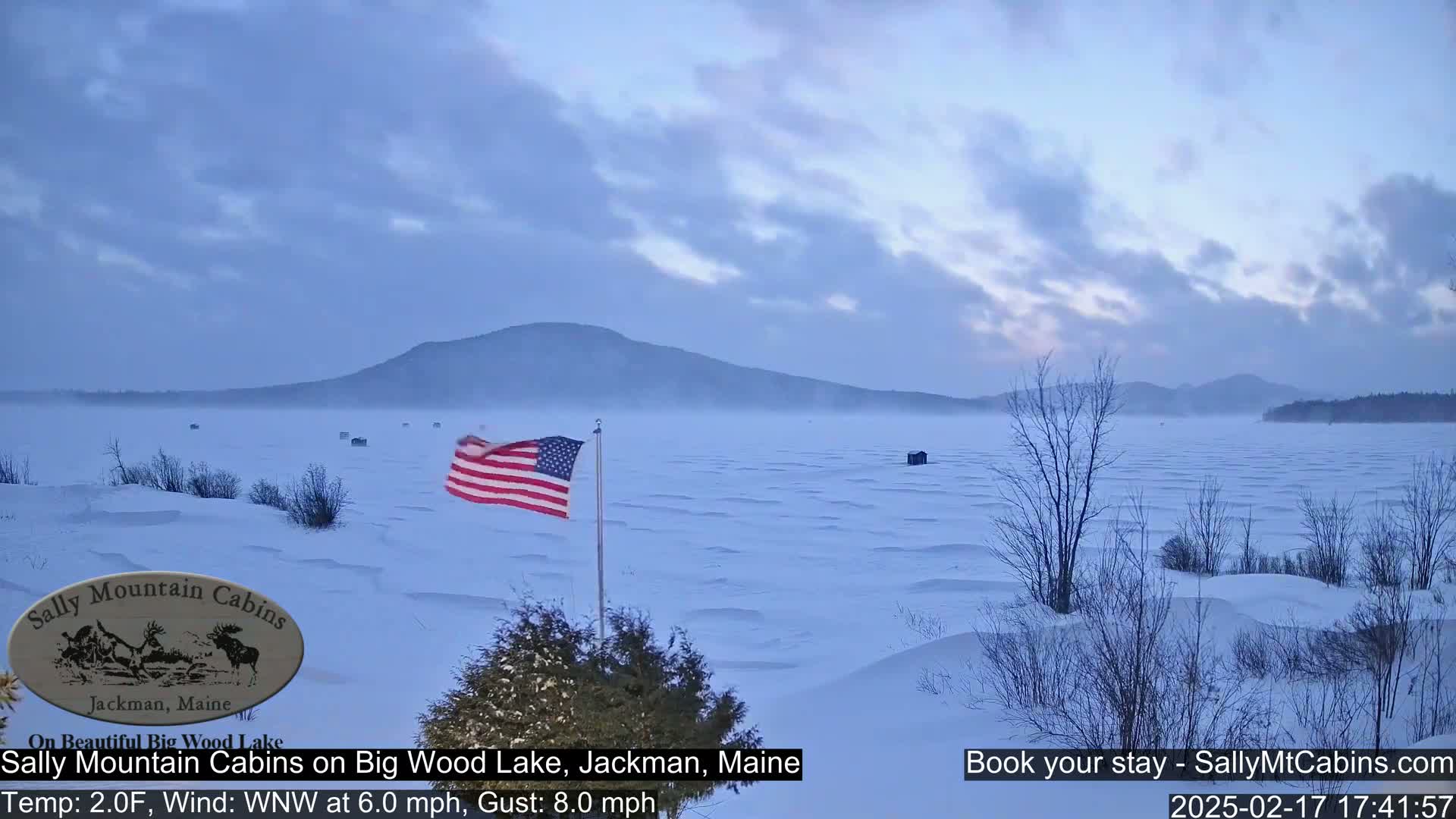 A snow-covered lake with a distant mountain range under a partly cloudy sky, a US flag waving in the wind.