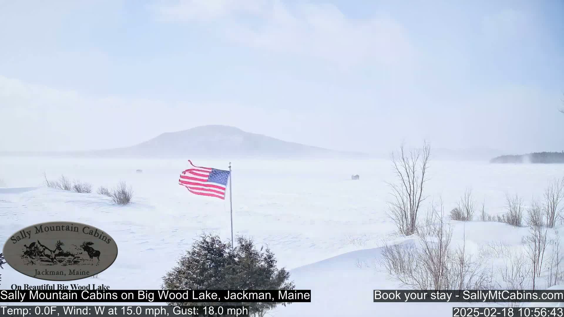 A snow-covered lake stretches to a hazy mountain under a light blue, windy sky, with an American flag waving in the foreground.