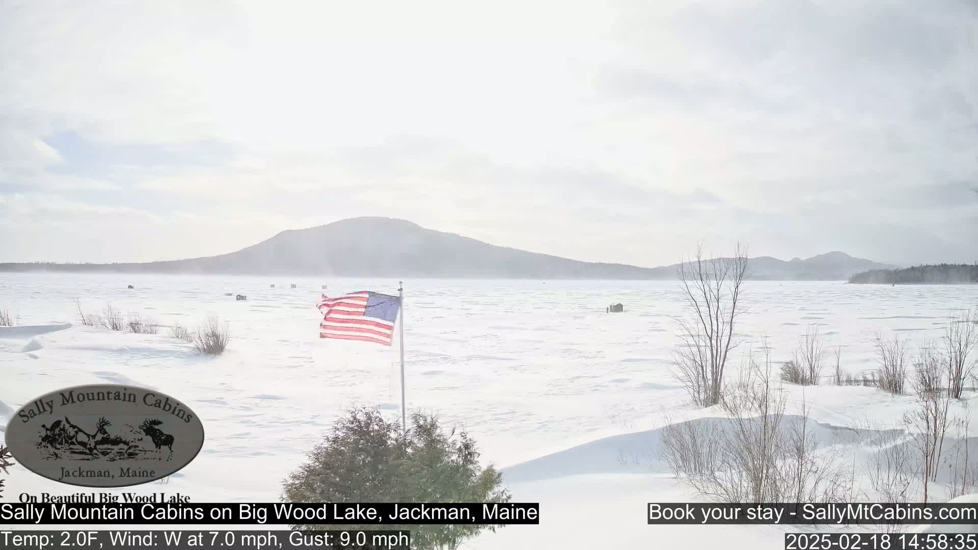 A snow-covered lake stretches to a distant mountain range under a hazy sky, with an American flag waving in a westerly wind.