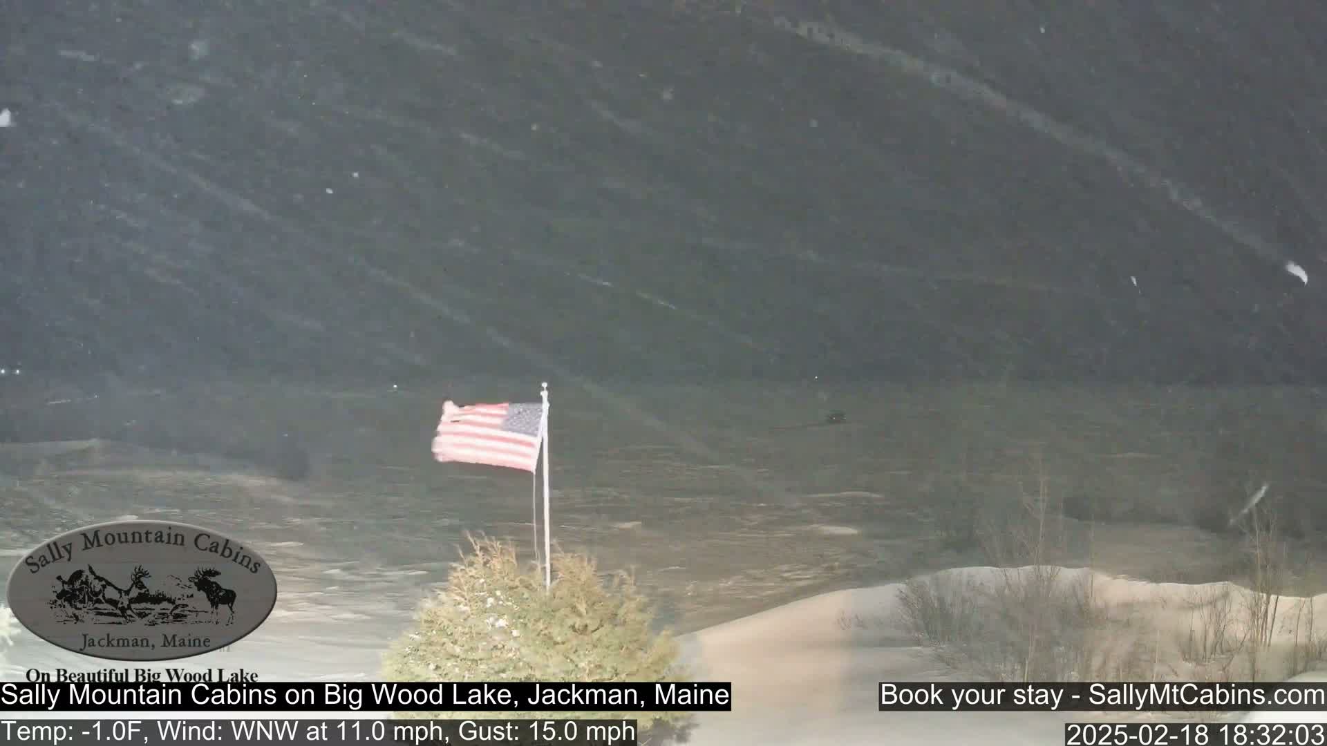 A partly snowy scene shows a body of water under a dark sky, with an American flag waving on a flagpole in the foreground.