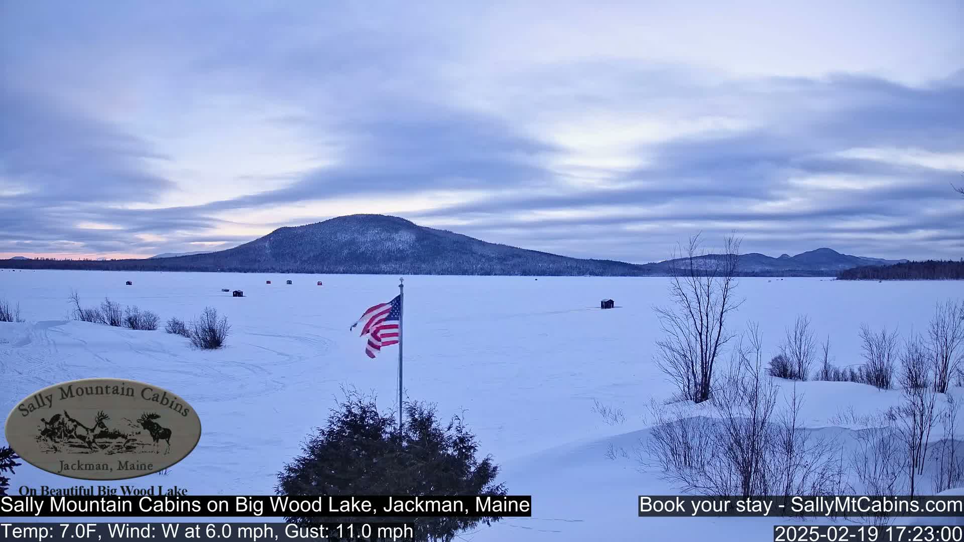 A frozen lake under a cloudy, twilight sky is viewed from a snowy shoreline, with a small American flag planted in the foreground and distant mountains in the background.
