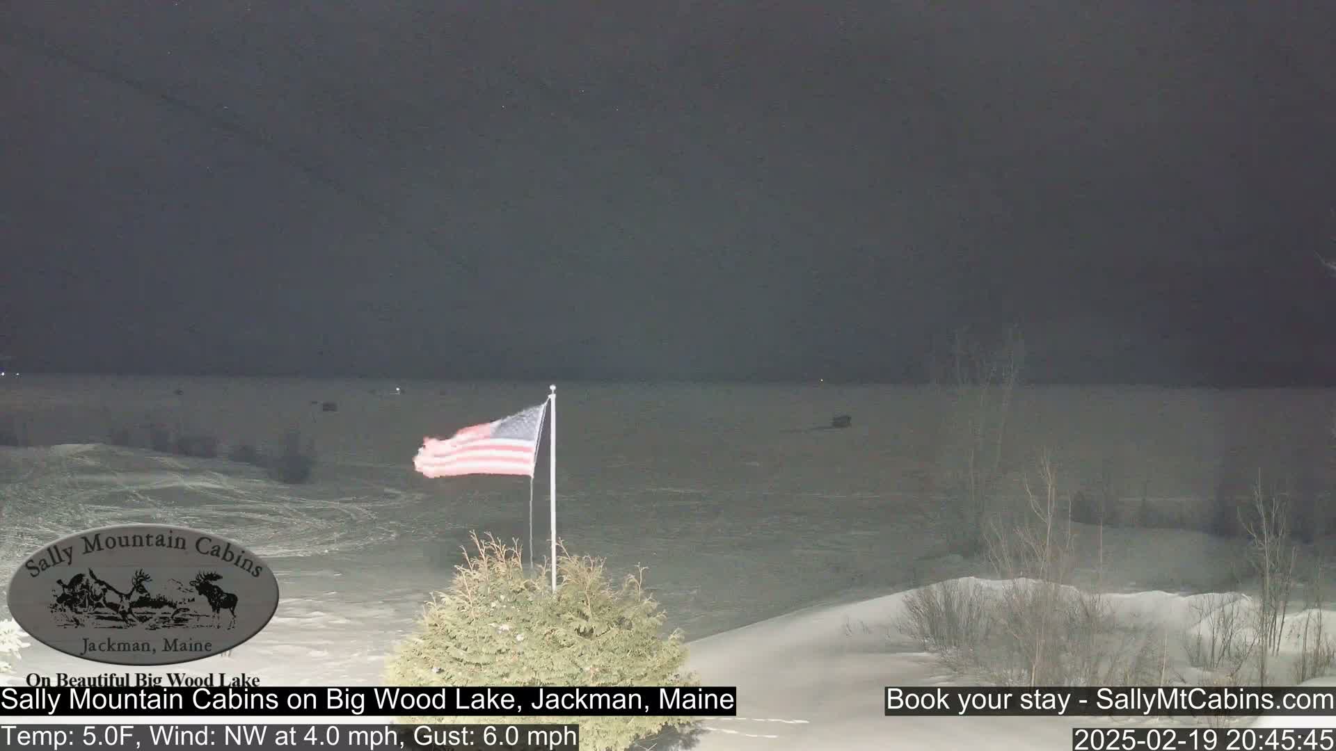 A nighttime view of a snow-covered lake under a dark sky, with an American flag waving in the foreground.