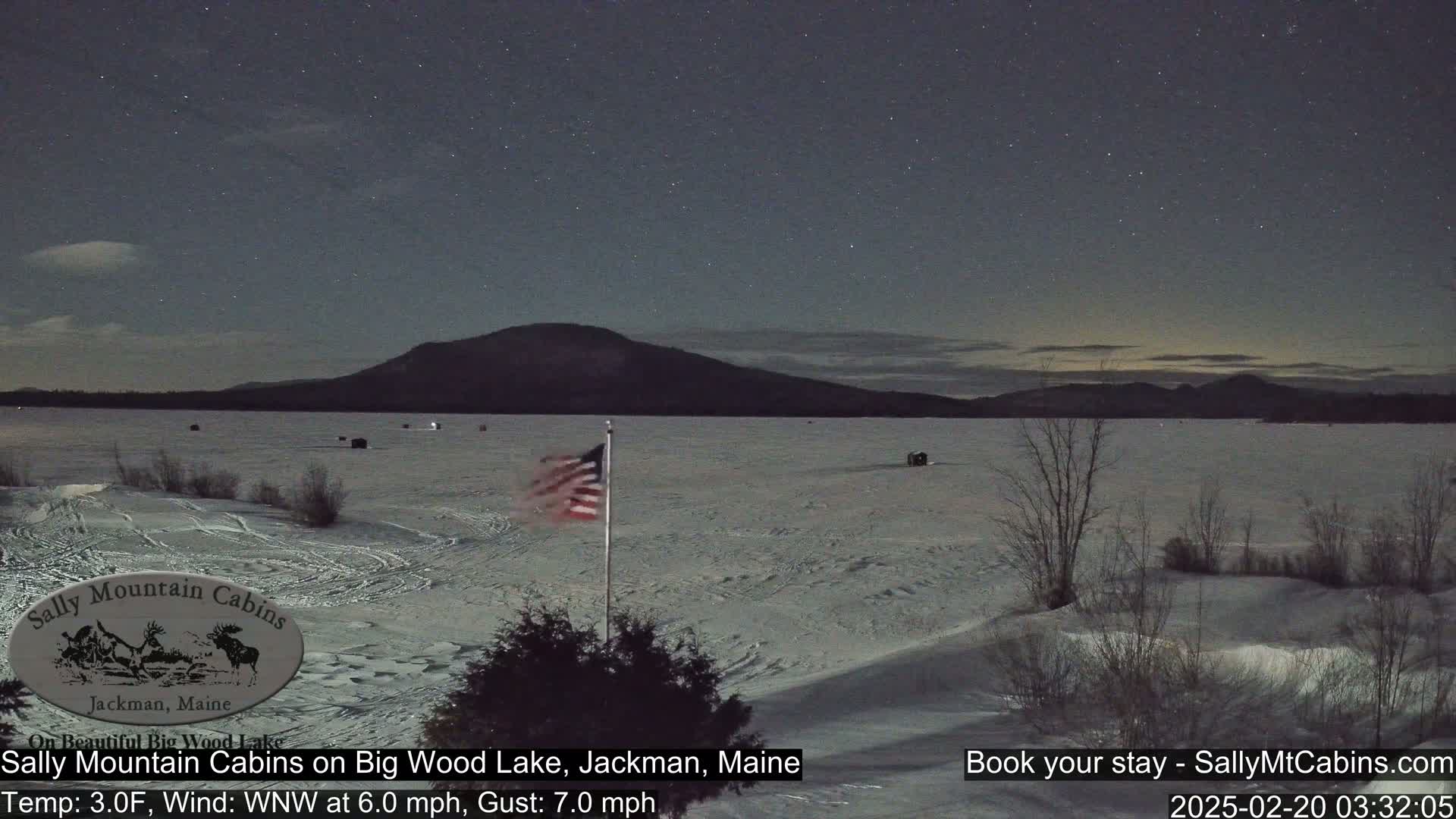 A snow-covered frozen lake under a starry night sky, with a distant mountain range and an American flag waving in the cold wind.