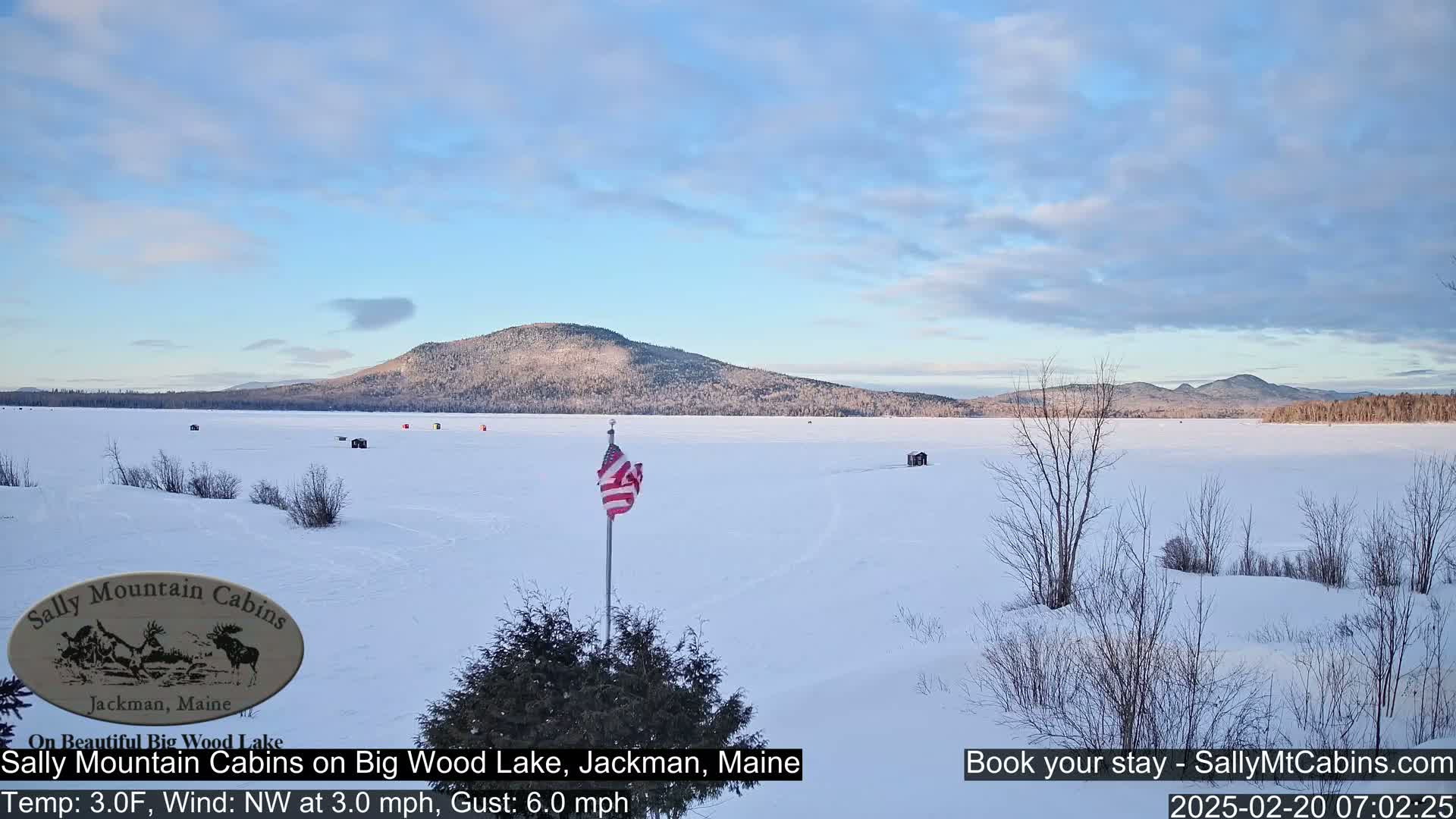 A snow-covered frozen lake stretches out before a line of distant, wooded hills under a partly cloudy sky.