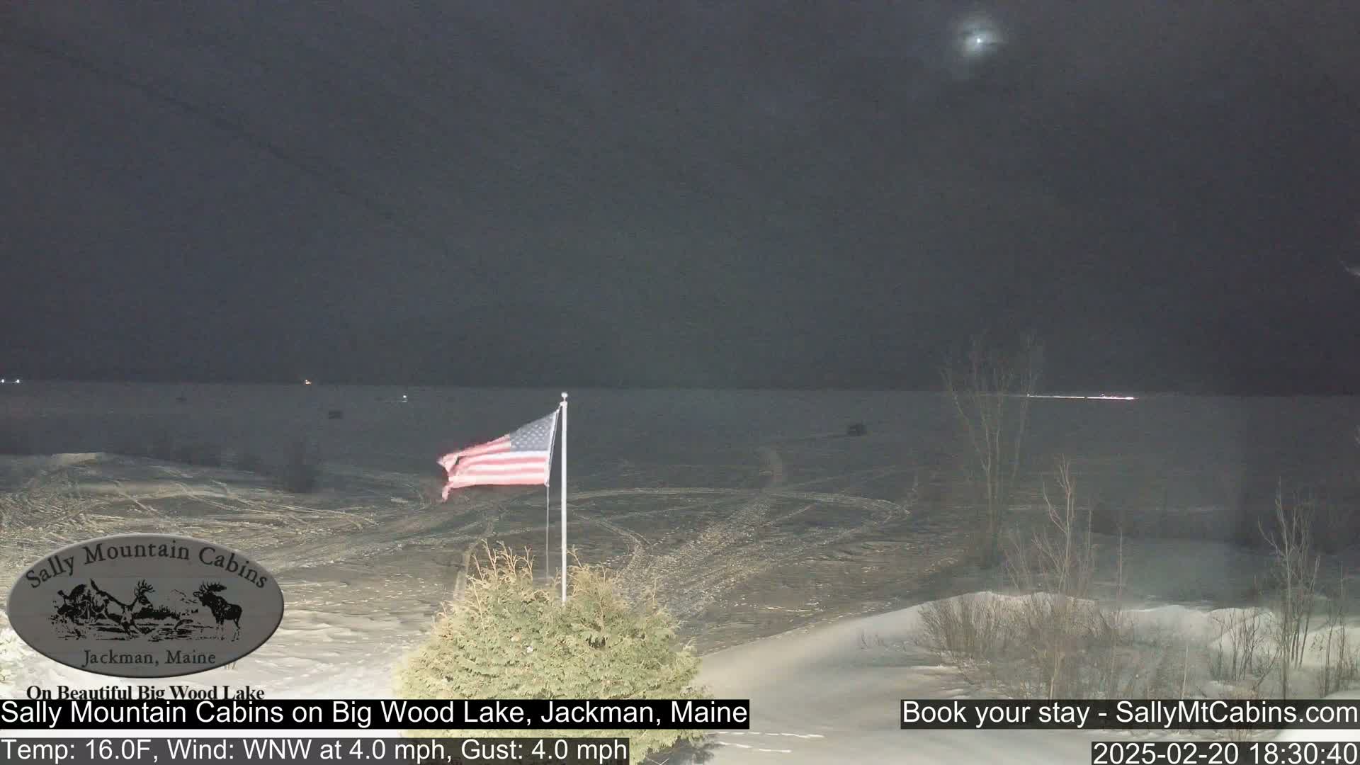 A nighttime view of a frozen lake with snow-covered banks, a waving American flag, and a few distant lights under a dark sky.