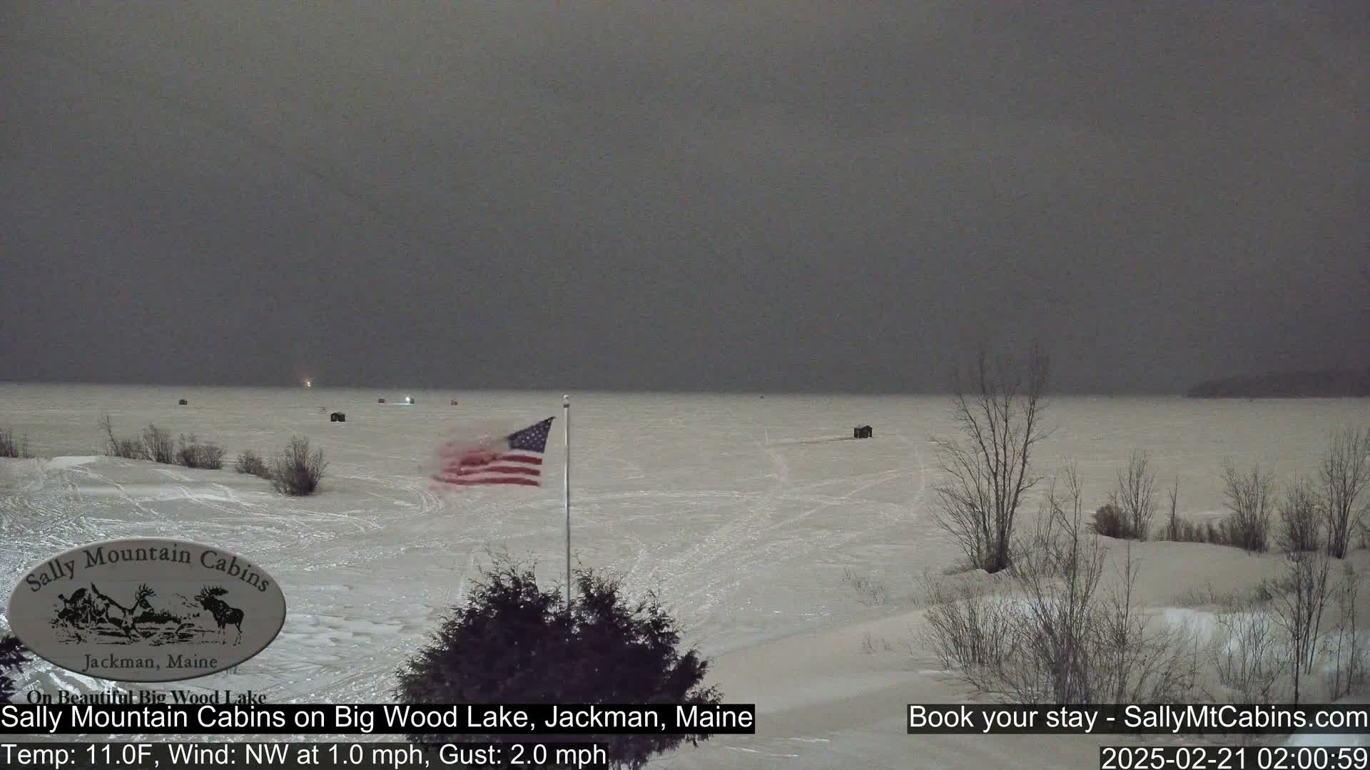 A snow-covered frozen lake is visible under a dark, overcast sky, with a few ice fishing huts and an American flag in the foreground.