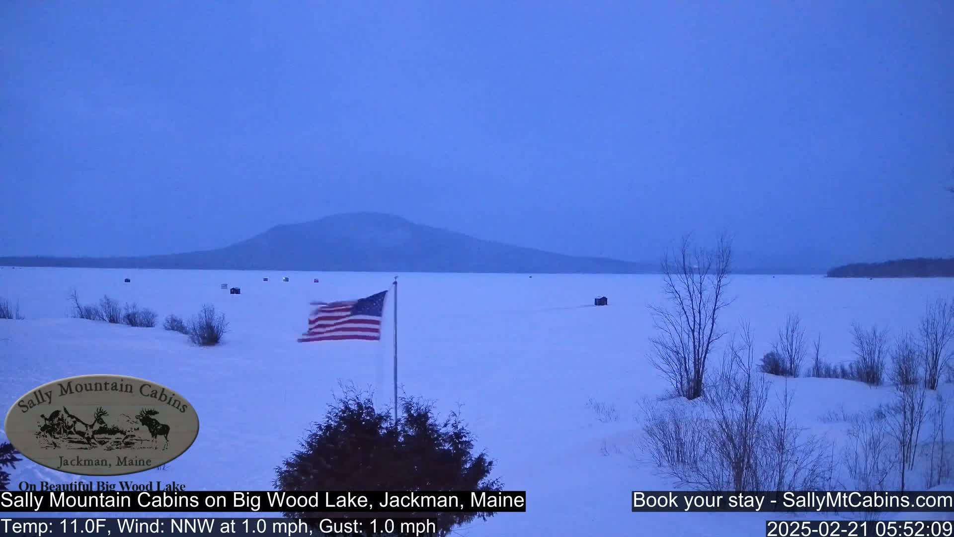 A snow-covered frozen lake stretches to a distant mountain under a twilight sky, with a few ice fishing shelters visible and an American flag waving in a light north-northwest wind.