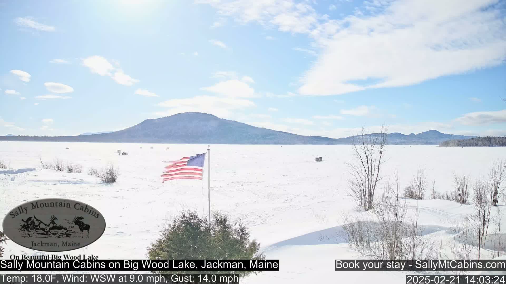 A snow-covered lake stretches to a distant mountain range under a partly cloudy sky, with a US flag fluttering in the breeze.