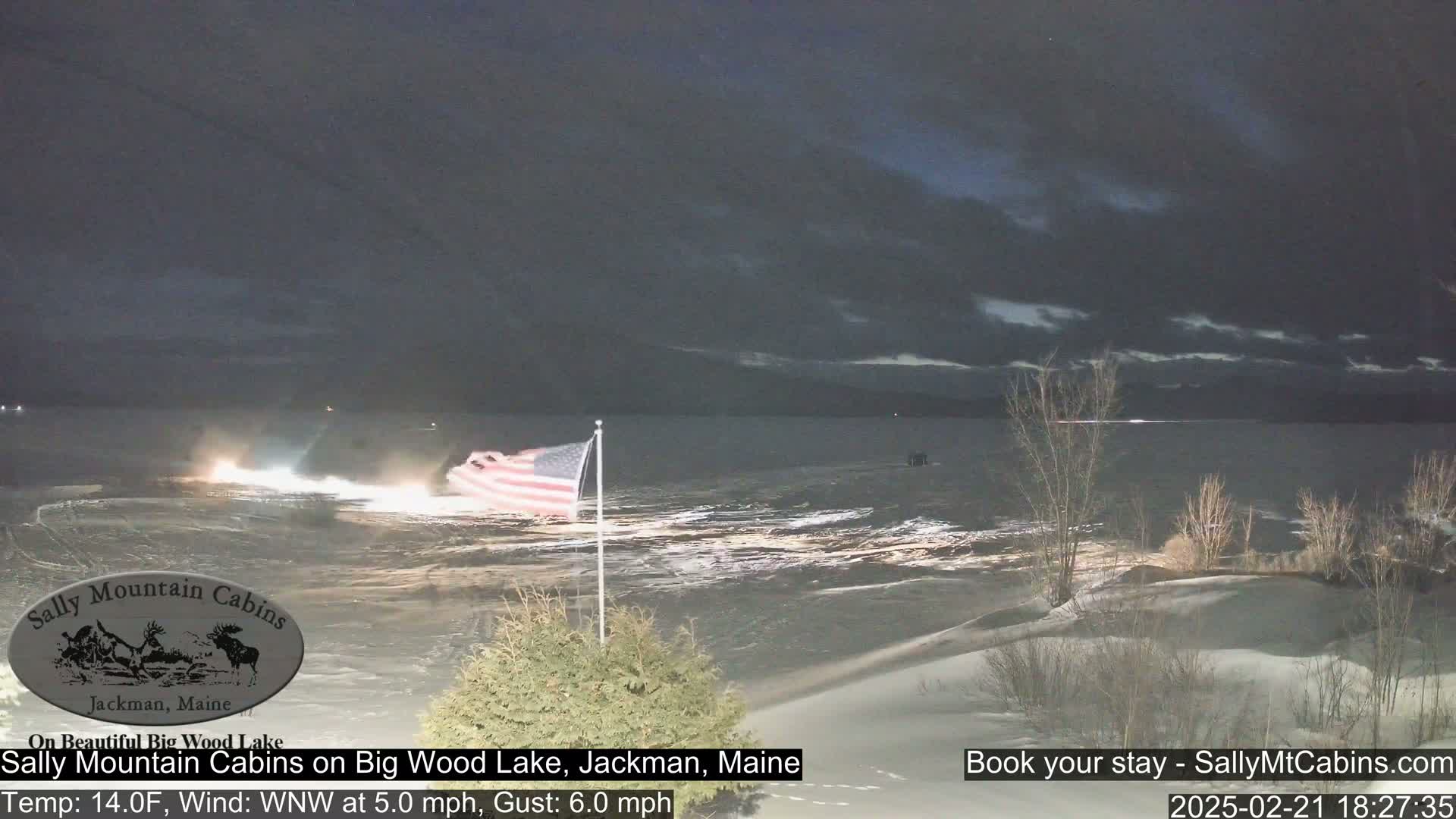 A nighttime view of a partially frozen lake with a gently waving American flag, under a cloudy sky.