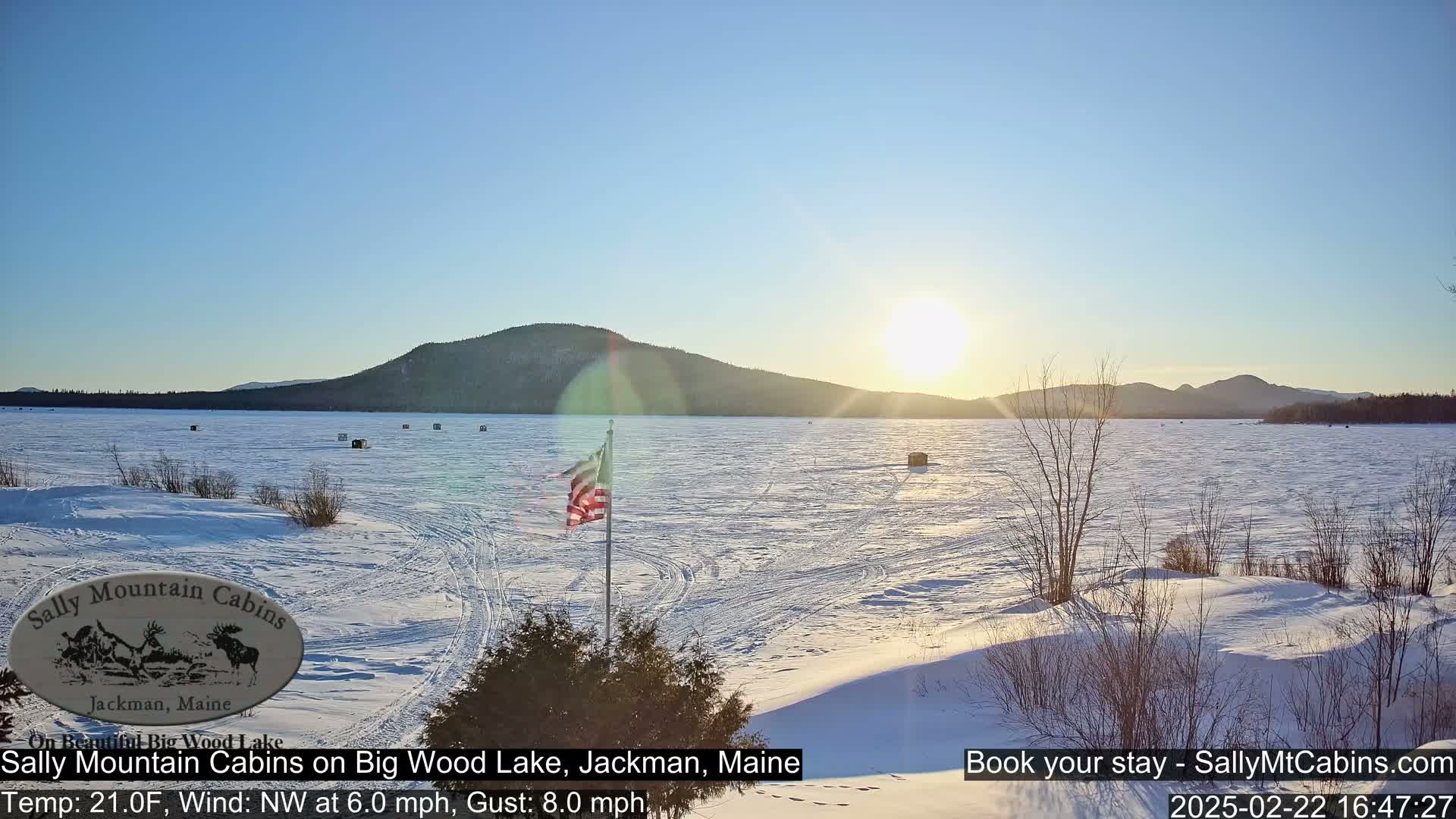 A frozen lake, under a clear, sunny sky, is seen from a snowy shoreline, with a distant mountain range and an American flag in the foreground.