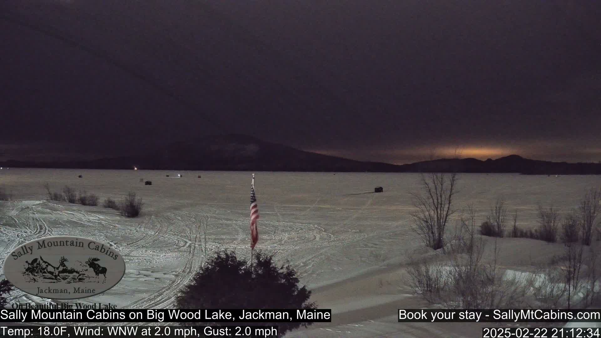 A frozen lake under a dark, twilight sky, with a few dark structures and a US flag visible near the shore.