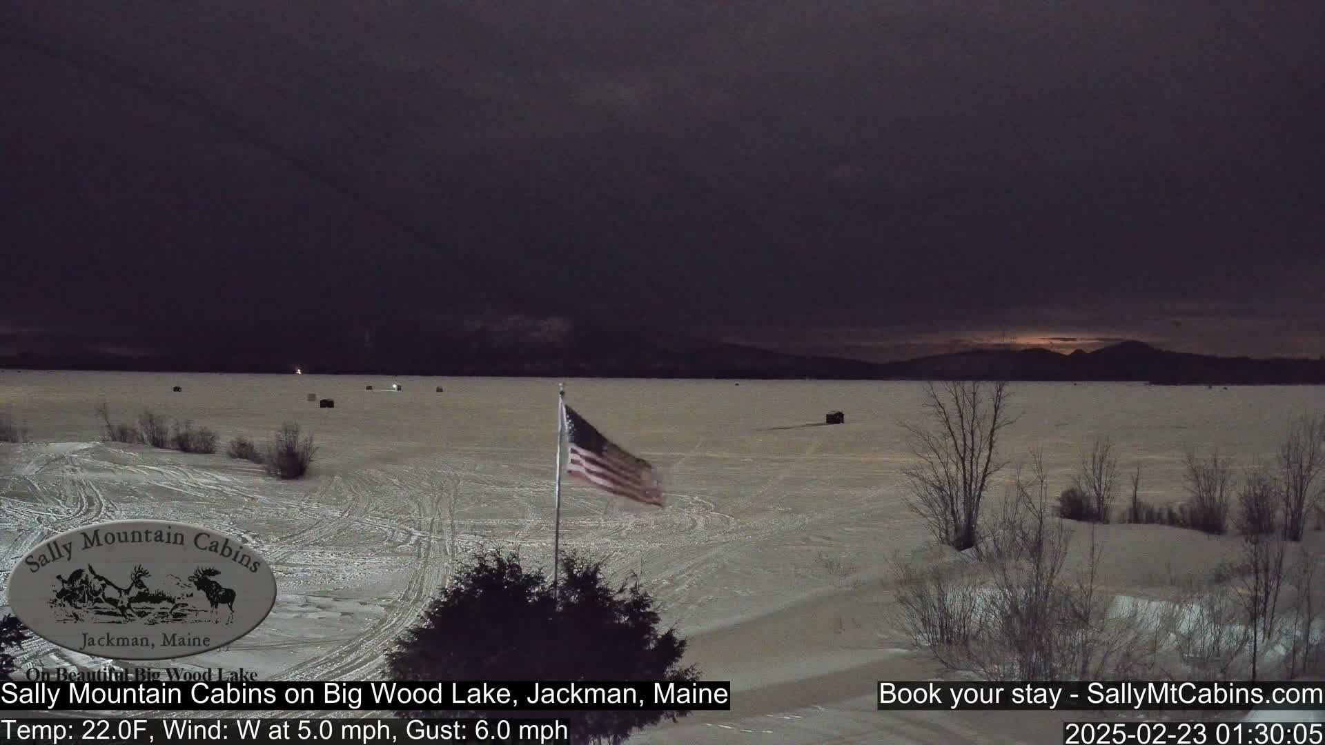 A frozen lake under a dark, overcast sky is viewed from a lakeside vantage point, with a small American flag waving in the wind.