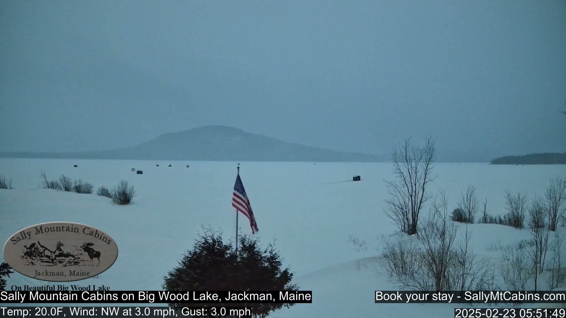 A snow-covered frozen lake stretches to a distant, hazy mountain range under an overcast sky, with a few ice fishing huts visible and an American flag standing in the foreground.