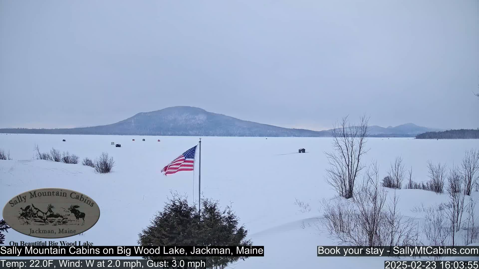 A snow-covered frozen lake stretches to a distant mountain range under an overcast sky, with a few ice fishing shelters visible and an American flag in the foreground.