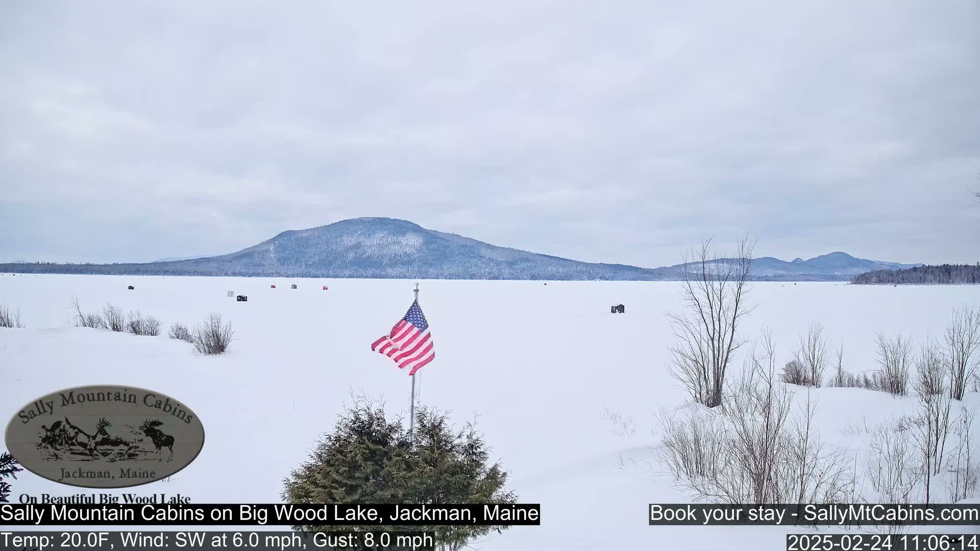 A snow-covered frozen lake stretches to a distant mountain range under a cloudy sky, with an American flag planted in the foreground.