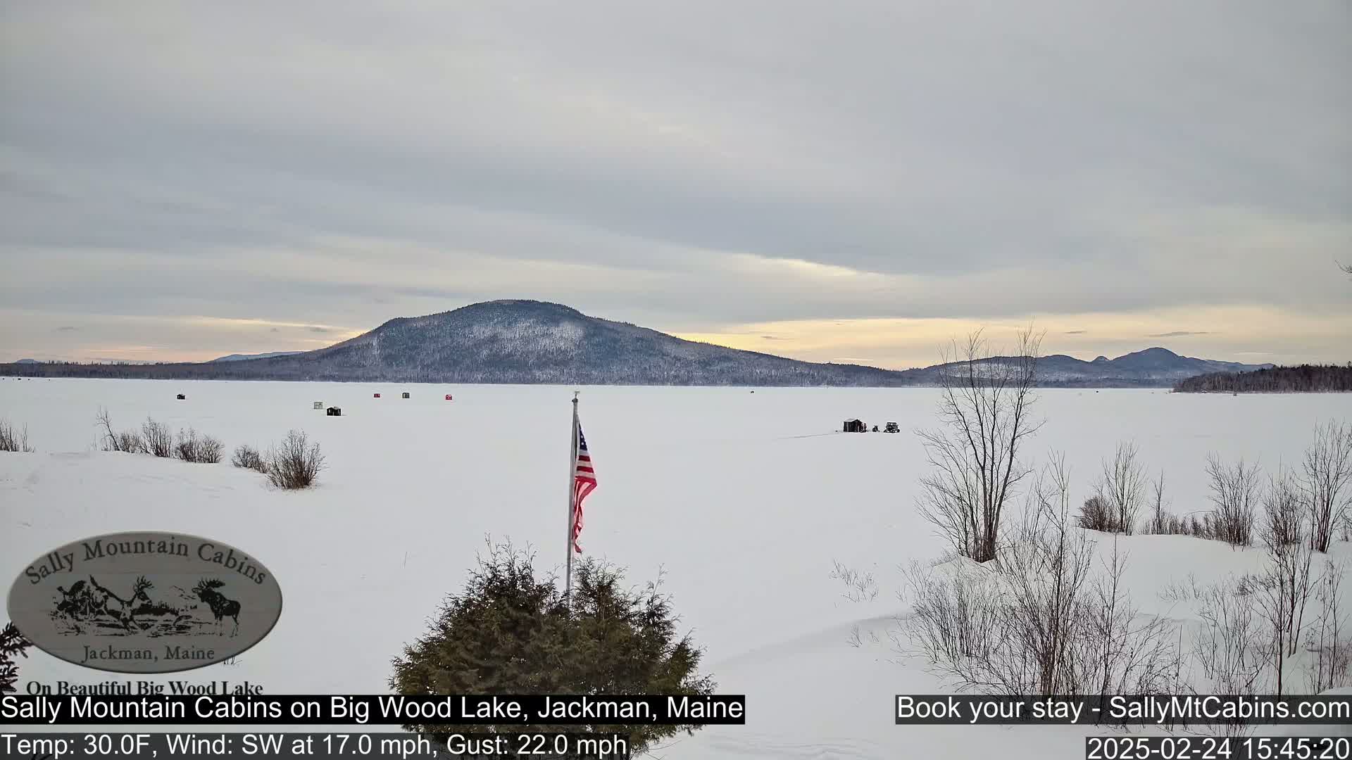 A frozen lake stretches toward snow-covered mountains under a mostly cloudy sky, with a few ice fishing shanties visible and an American flag standing near the foreground.