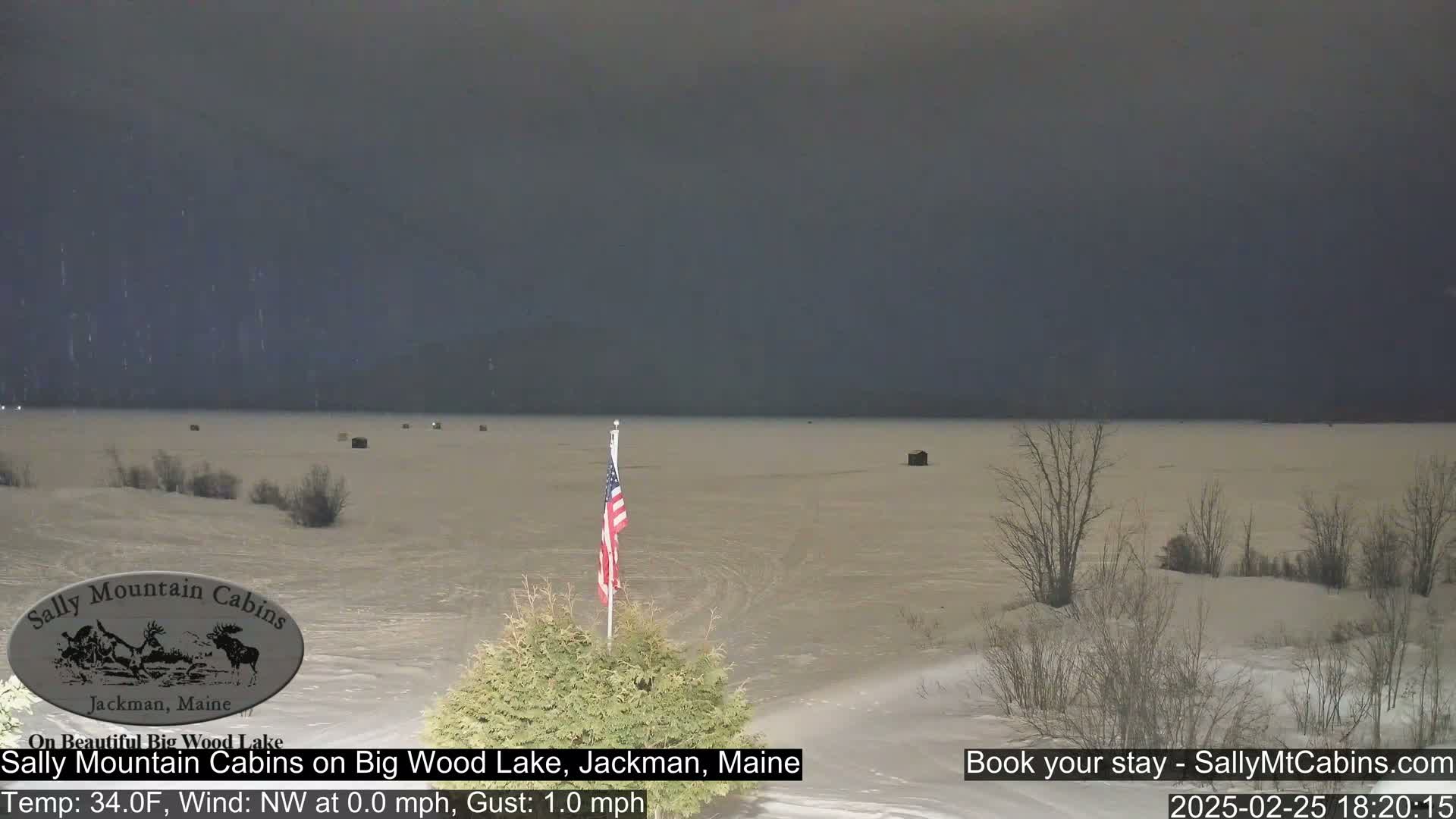 A mostly frozen lake is visible under a dark, overcast sky, with a few small structures and bare trees along the shoreline, and an American flag in the foreground.