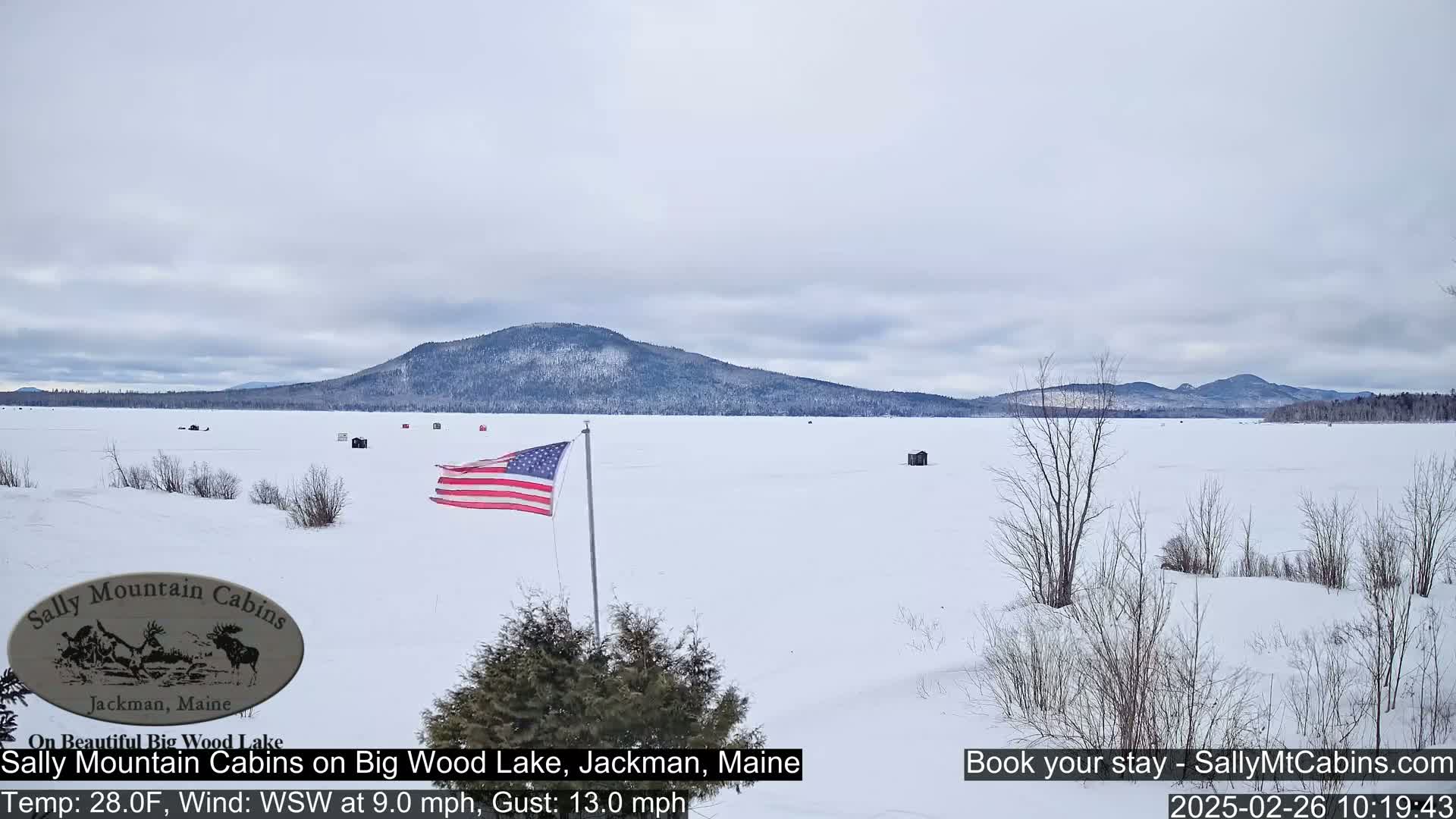 A snow-covered frozen lake stretches to a distant mountain range under a cloudy sky, with an American flag flying in the foreground.