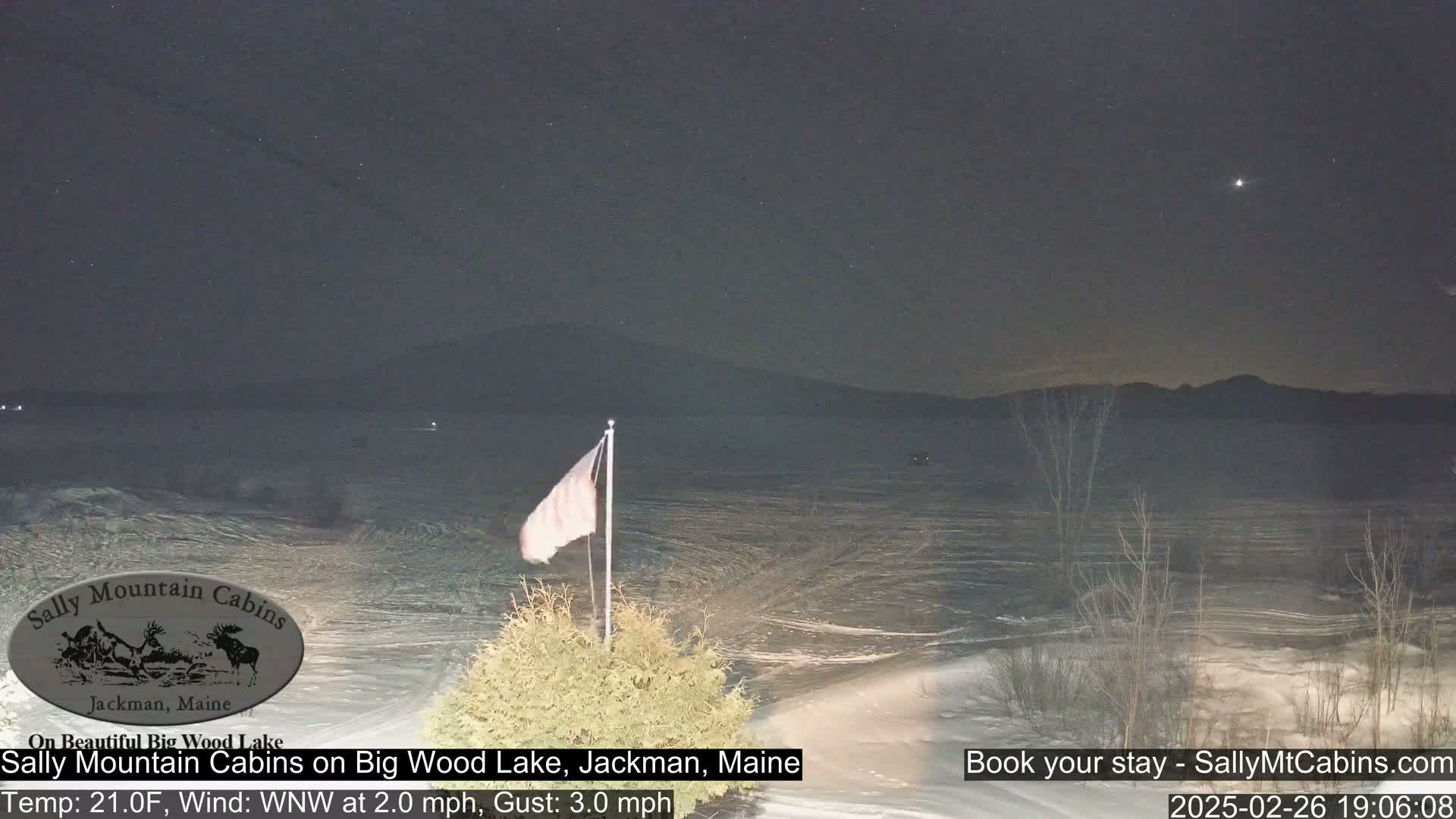 A nighttime view of a snow-covered lake under a mostly clear sky with a faint star visible, a flagpole with a flag, and some bare trees in the foreground.